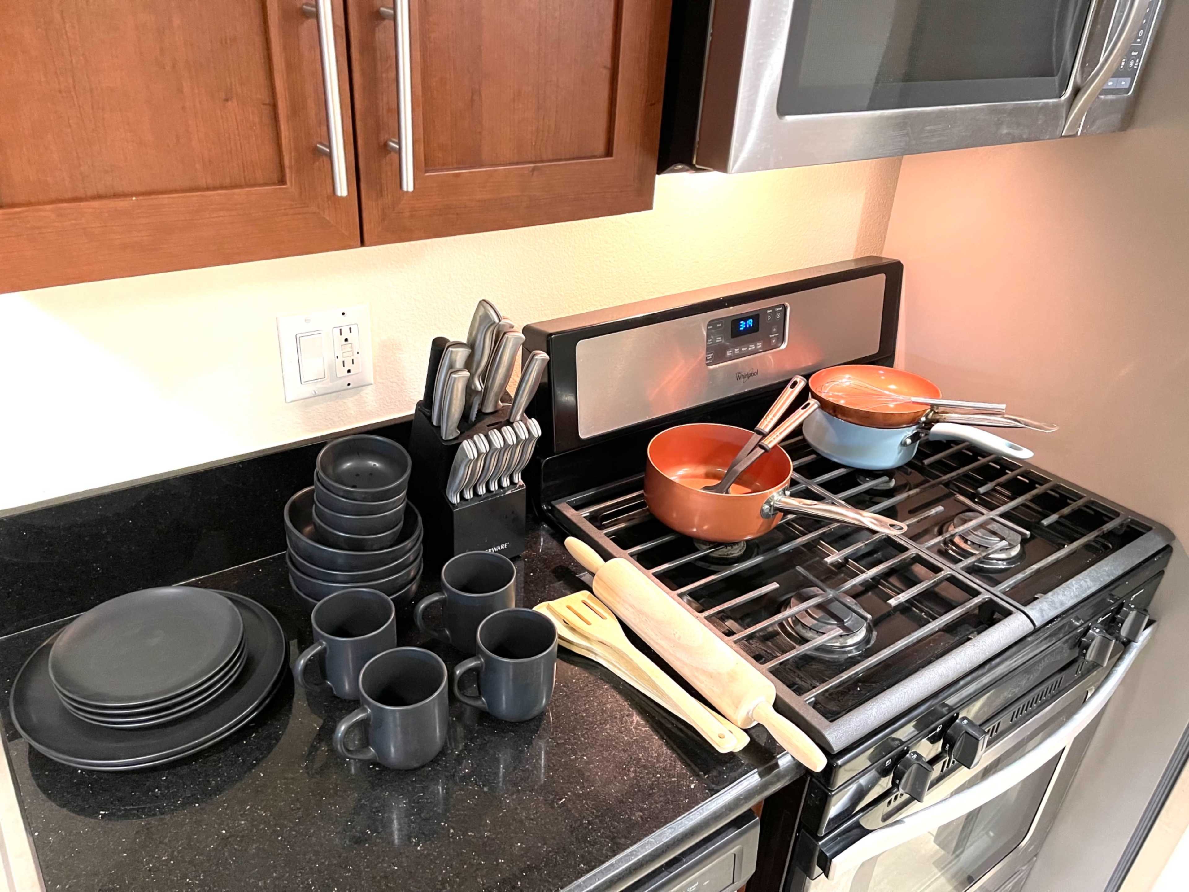 A kitchen countertop features a set of stacked black plates and mugs alongside pots, utensils, and a rolling pin on a gas stove.