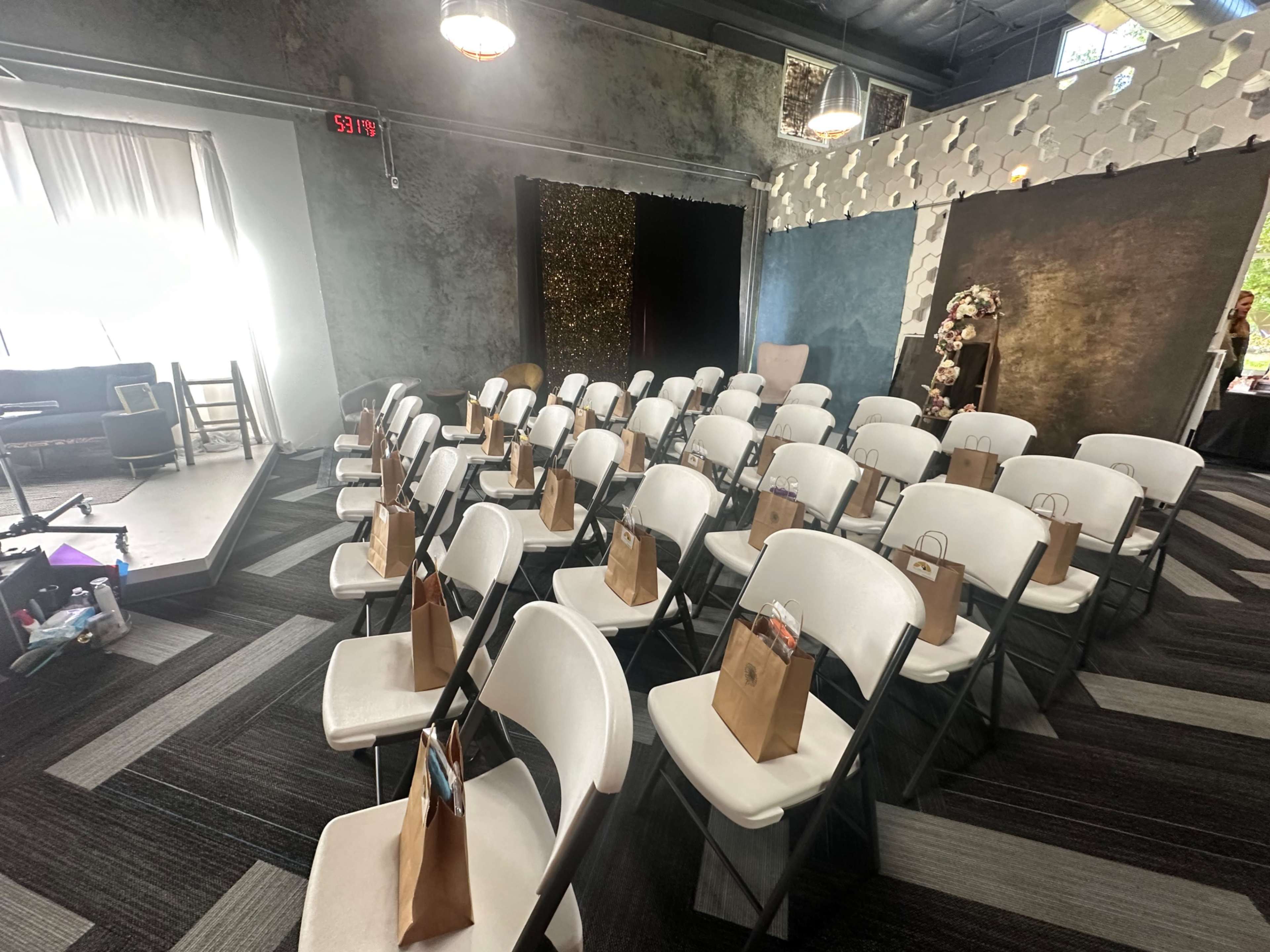 A row of white folding chairs is arranged in a spacious venue, each with a small gift bag resting on the seat.