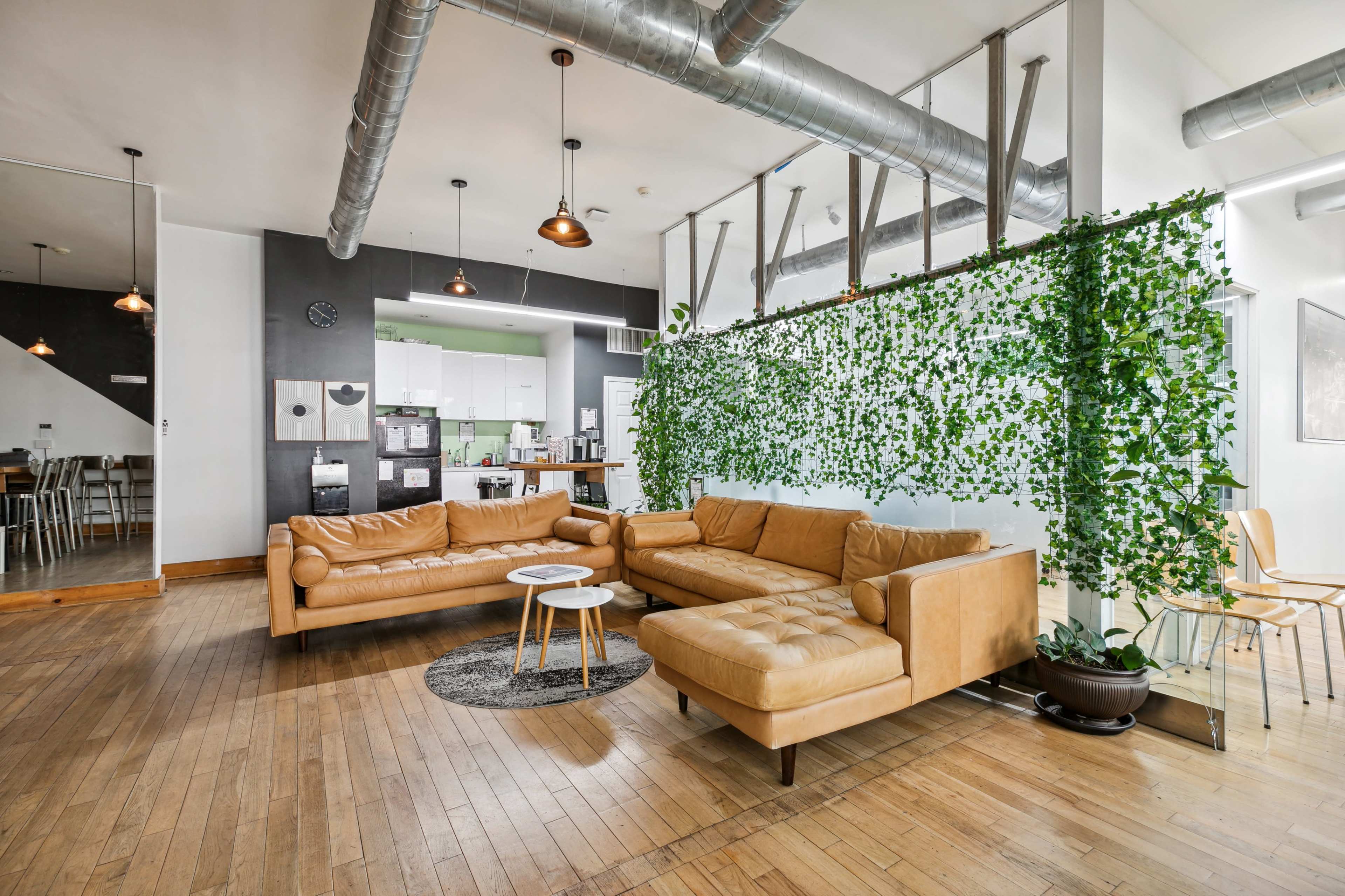 The image shows a modern living space featuring a tan sectional sofa, a small round table, and a wall adorned with green plants, with a kitchen area visible in the background.