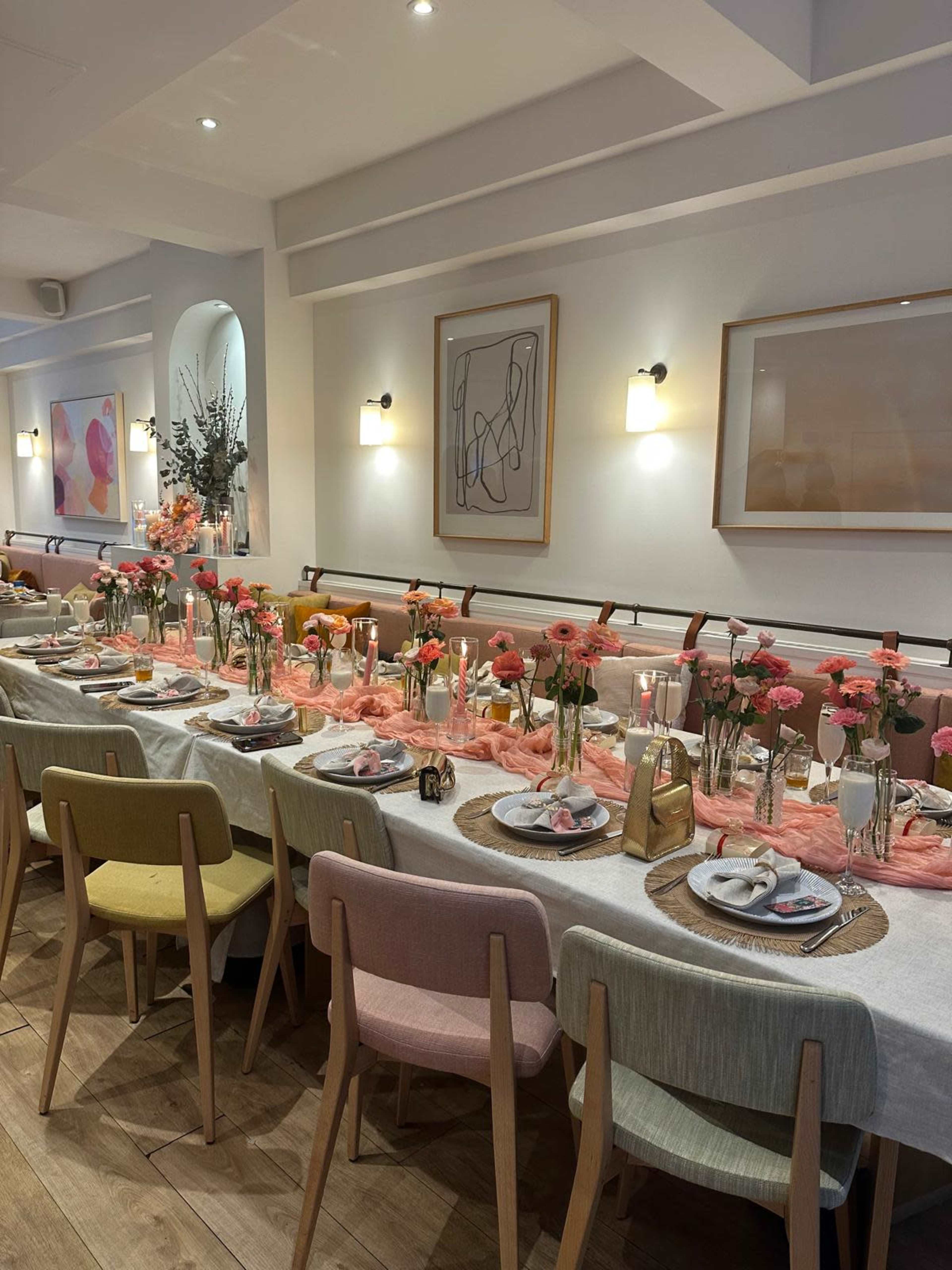 The image shows a long dining table set with floral centerpieces, place settings, and colorful chairs in a modern restaurant setting.