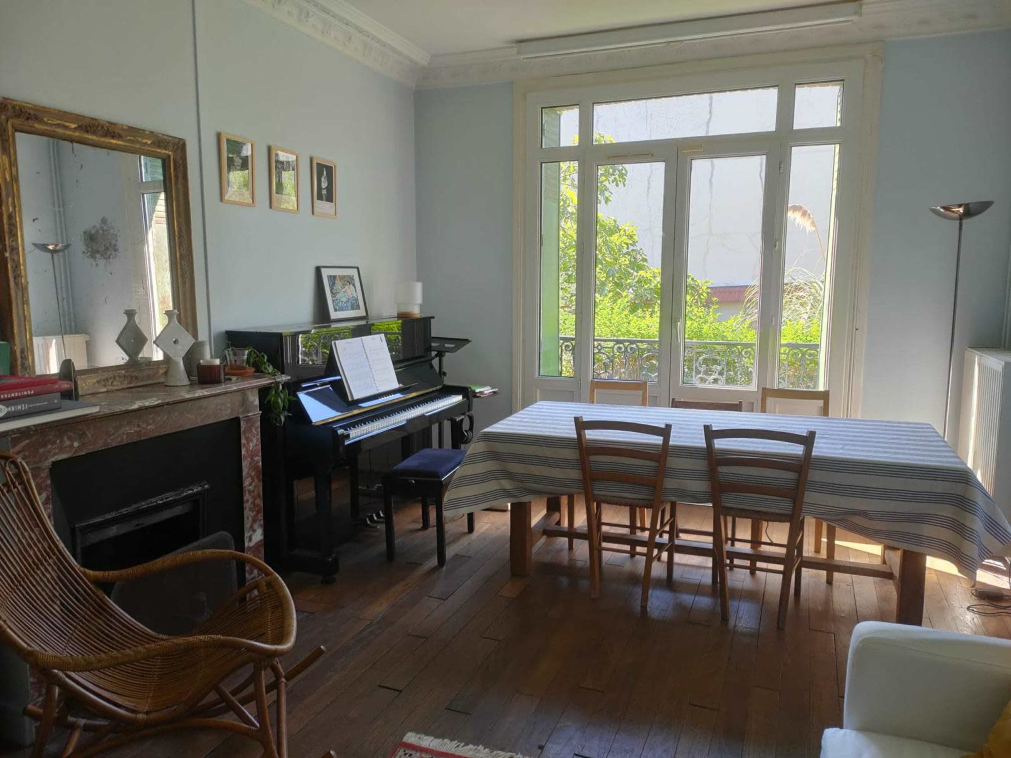 The room features a large dining table surrounded by chairs, a piano, and large windows allowing natural light to illuminate the space.