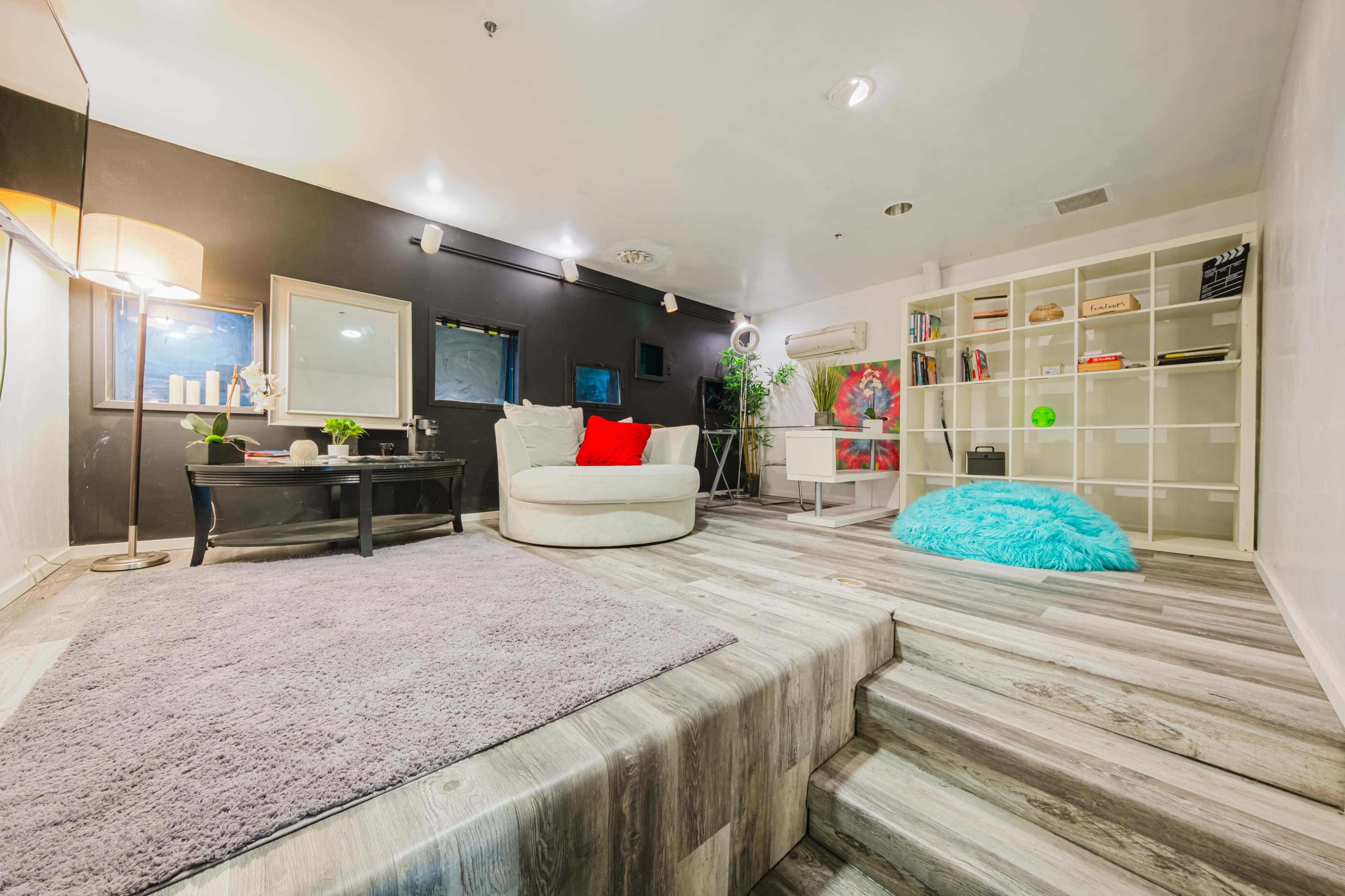 The image shows a modern lounge area with a circular armchair, a decorative rug, and shelves filled with various items, all set against a dark wall.