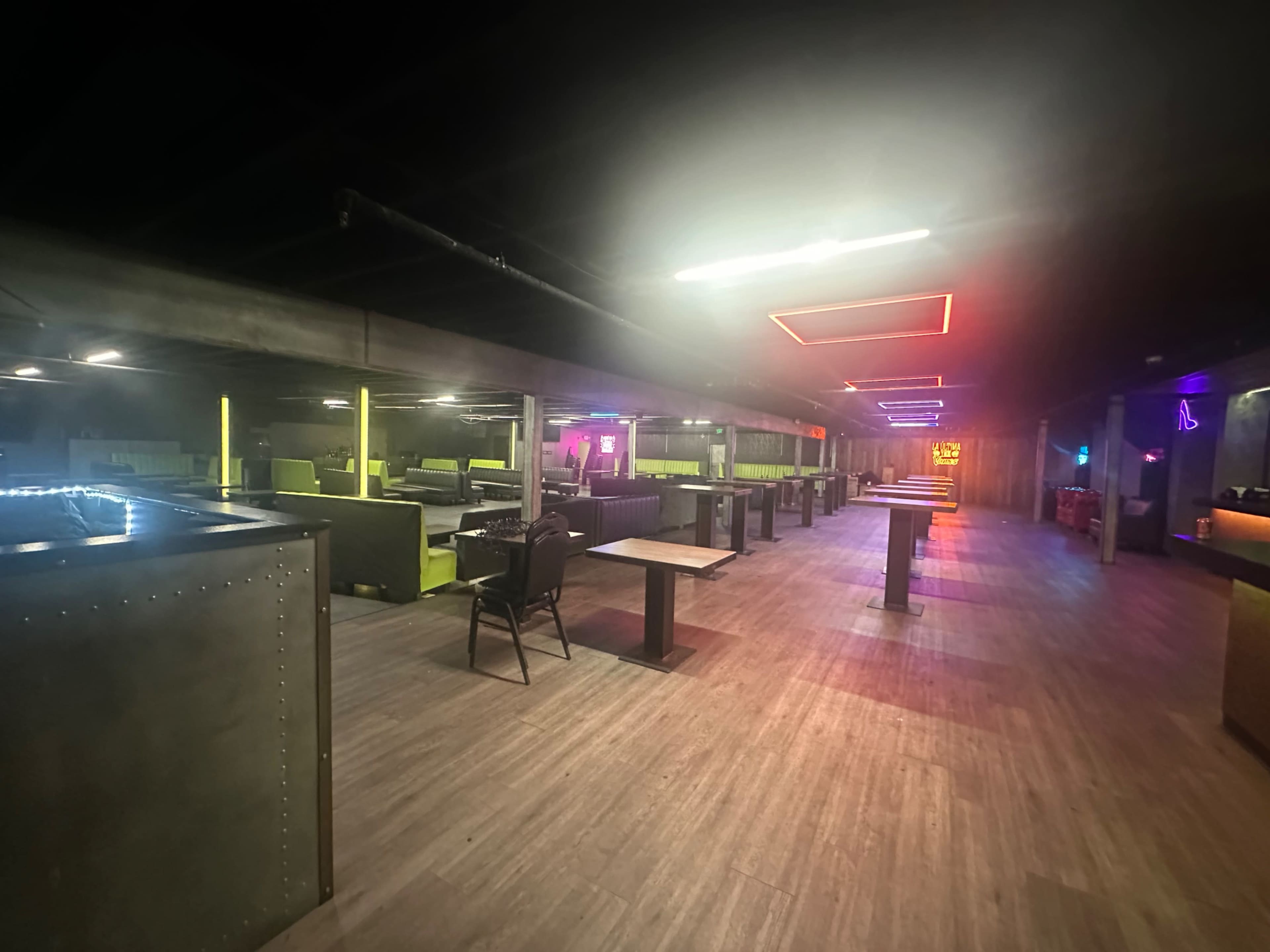 The image shows an empty, dimly lit interior of a lounge featuring tables, a bar area, and colorful neon lighting.