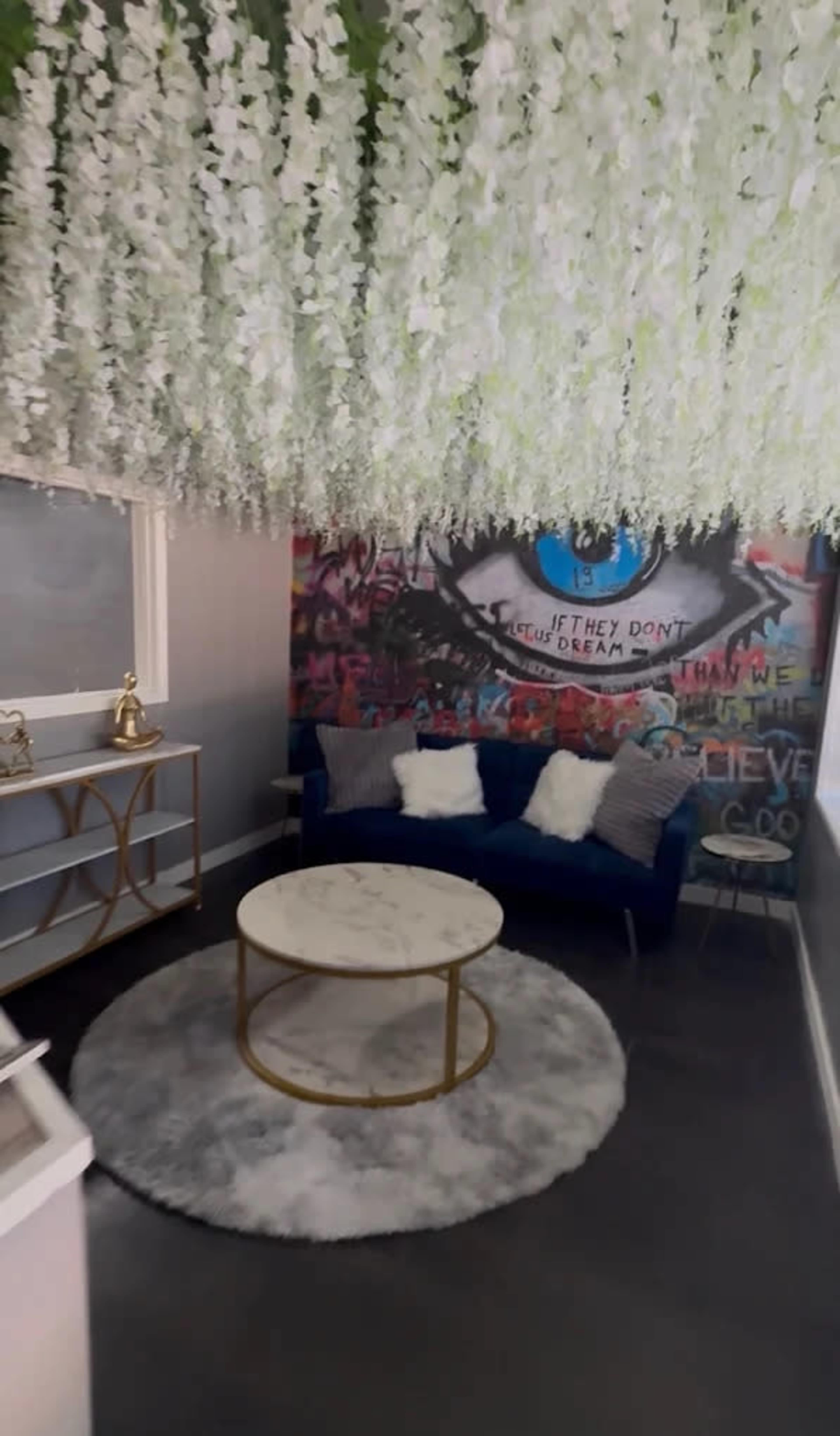 The image features a cozy interior space with a blue couch adorned with pillows, a circular marble coffee table, and an eye-catching floral ceiling, alongside a colorful mural on the wall.