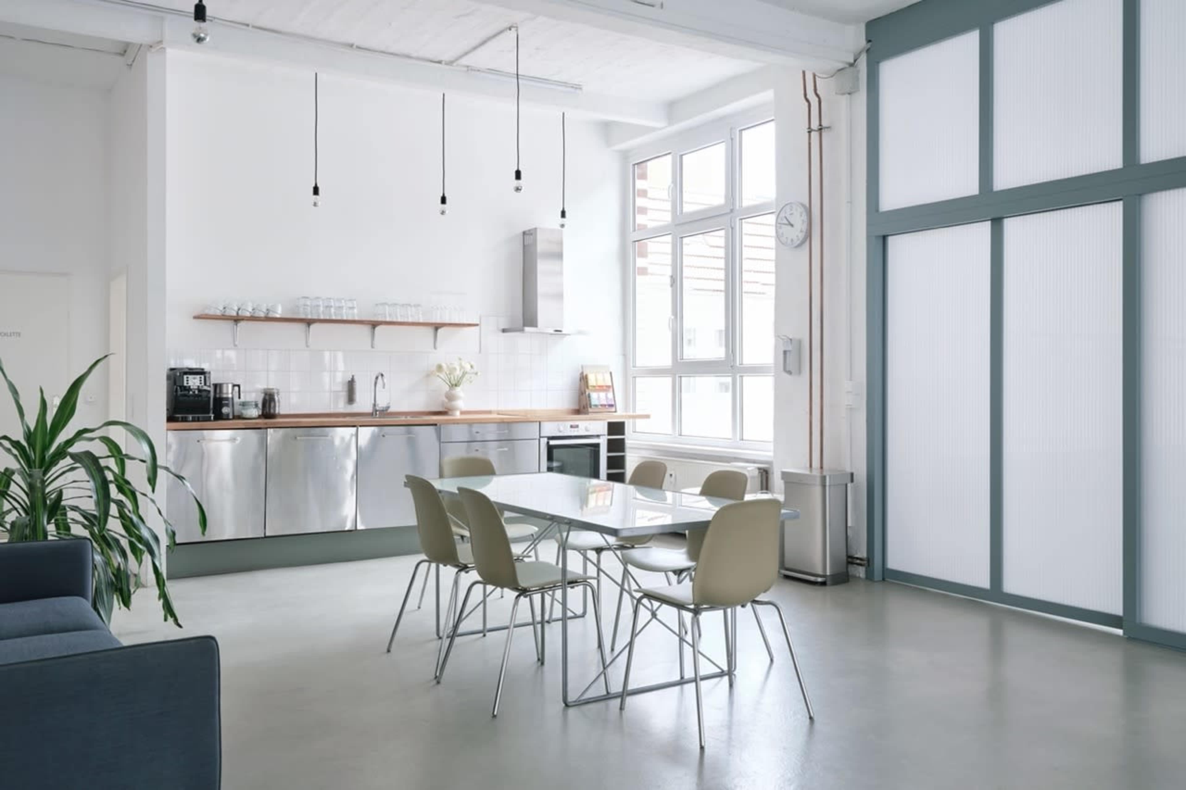 The image shows a bright, modern kitchen and dining area with a gray sofa, a sleek table surrounded by chairs, and large windows allowing natural light to fill the space.