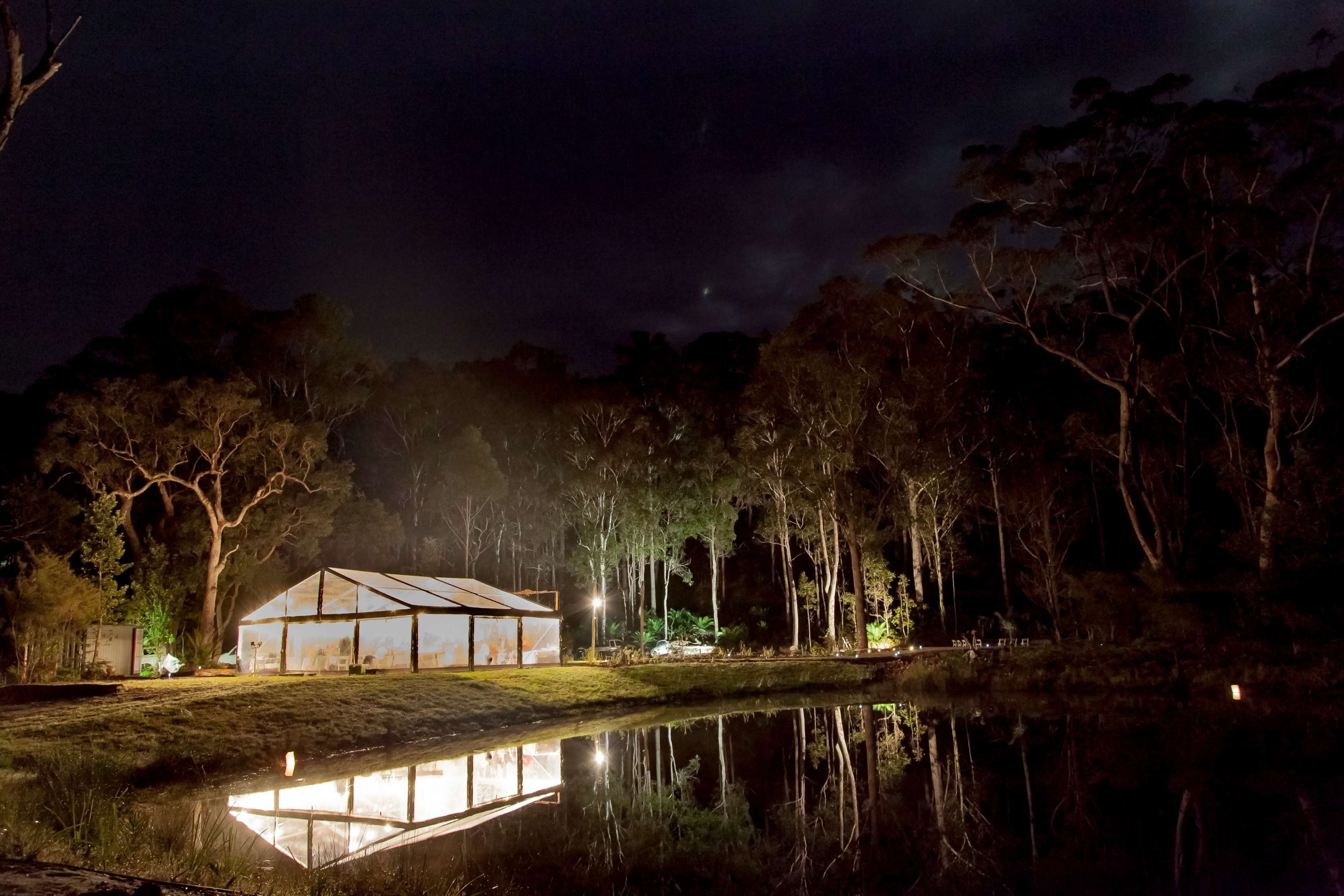 A white tent is illuminated at night beside a calm pond, surrounded by dense trees.