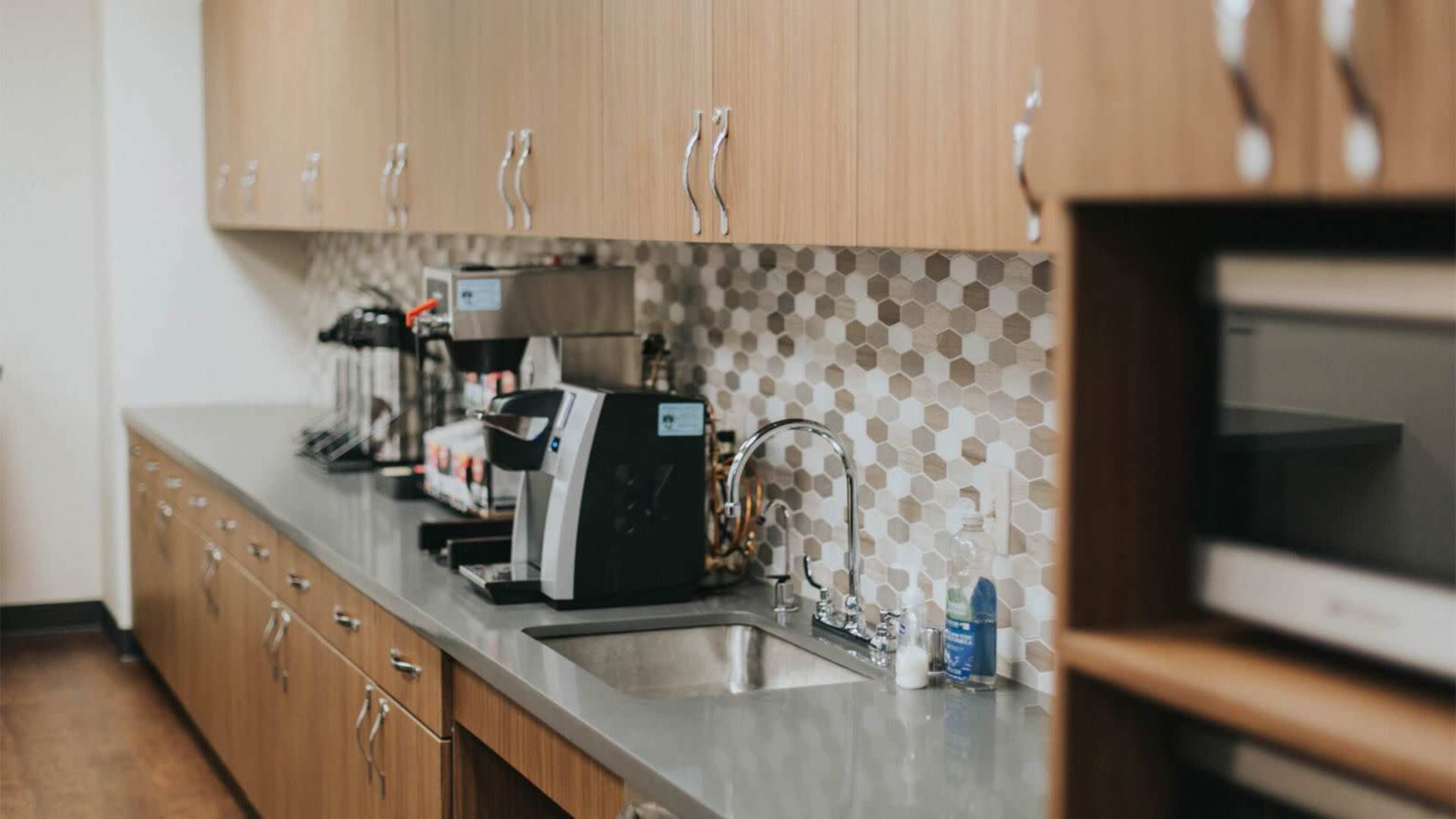 A modern kitchen area with a countertop, a sink, coffee machines, and a patterned backsplash.