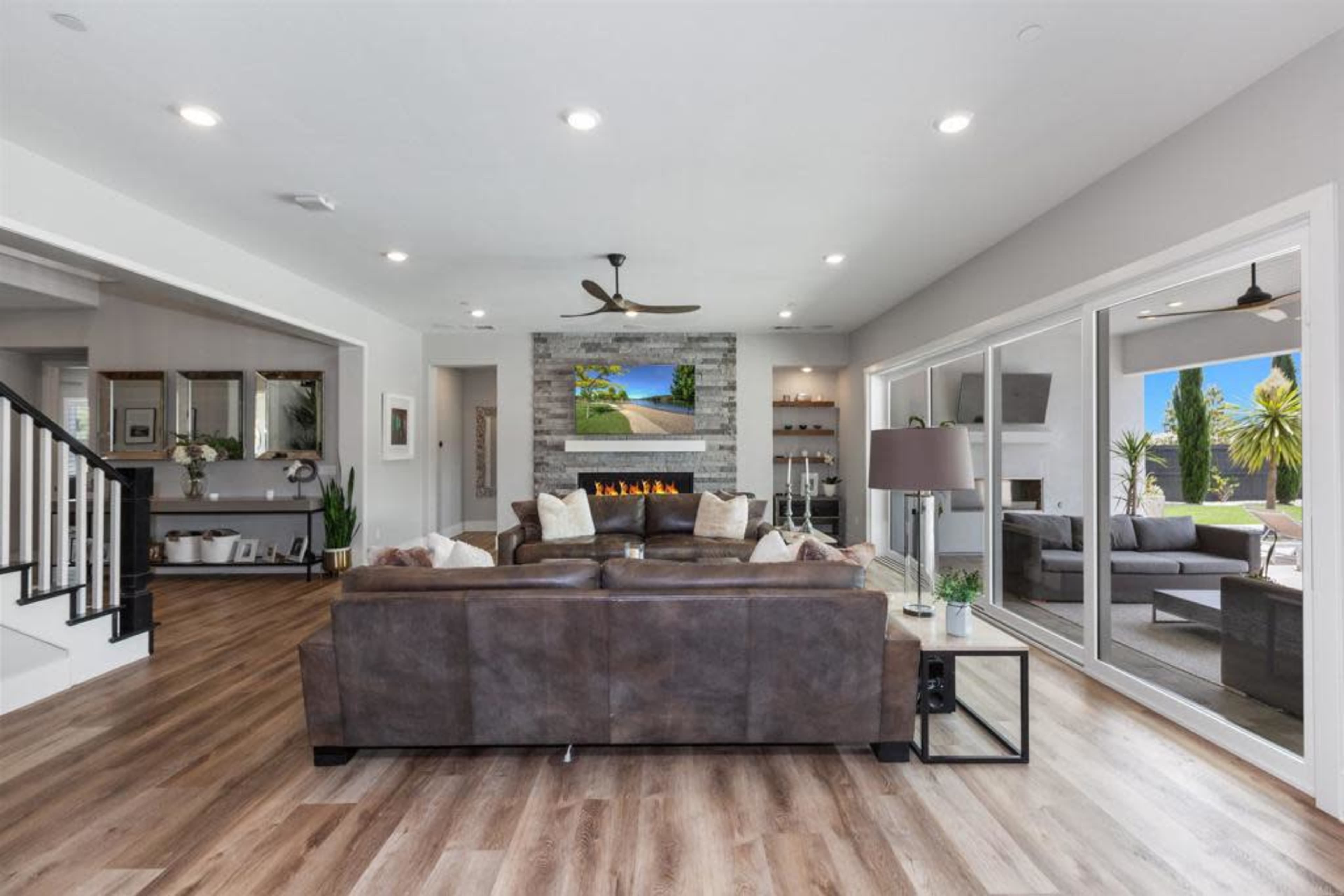 A modern living room with a brown leather sectional sofa, a stone fireplace, and large windows overlooking a patio with greenery.