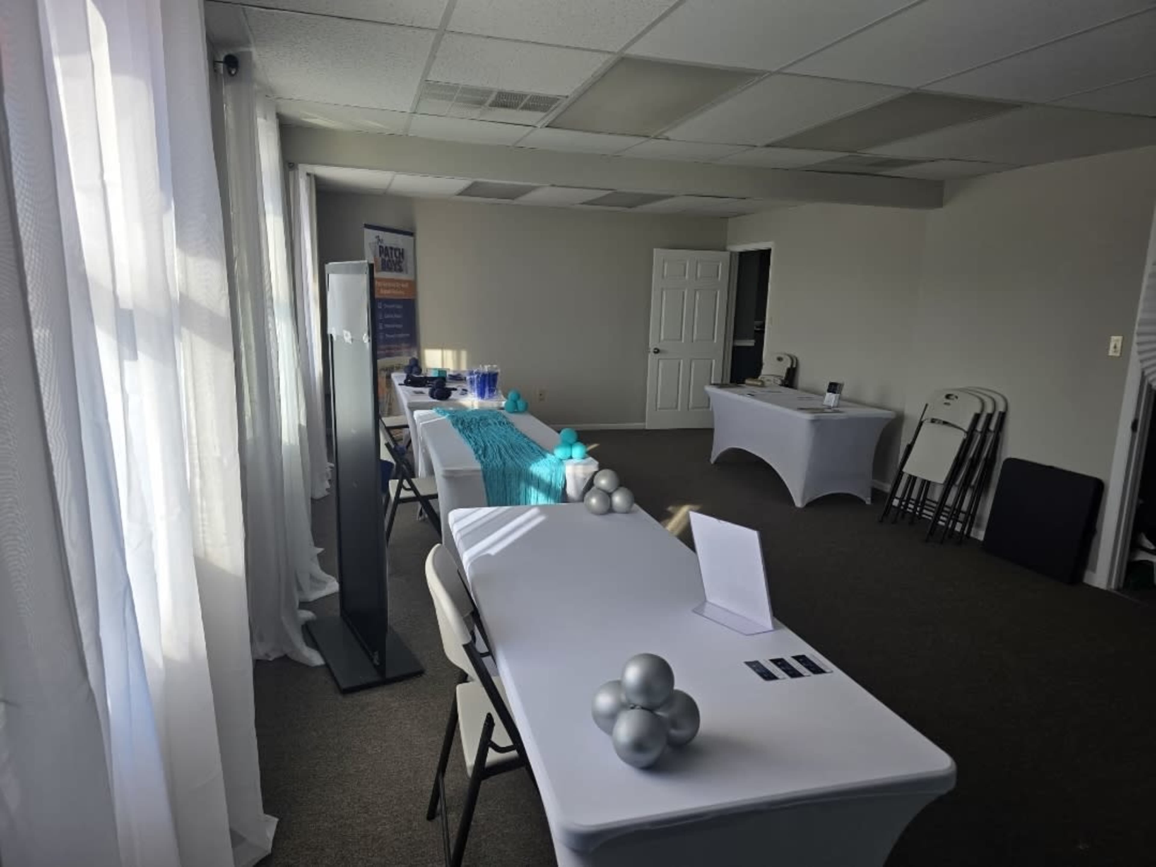 The image shows an empty event space with two tables dressed in white and teal decorations, along with chairs and a banner in the background.
