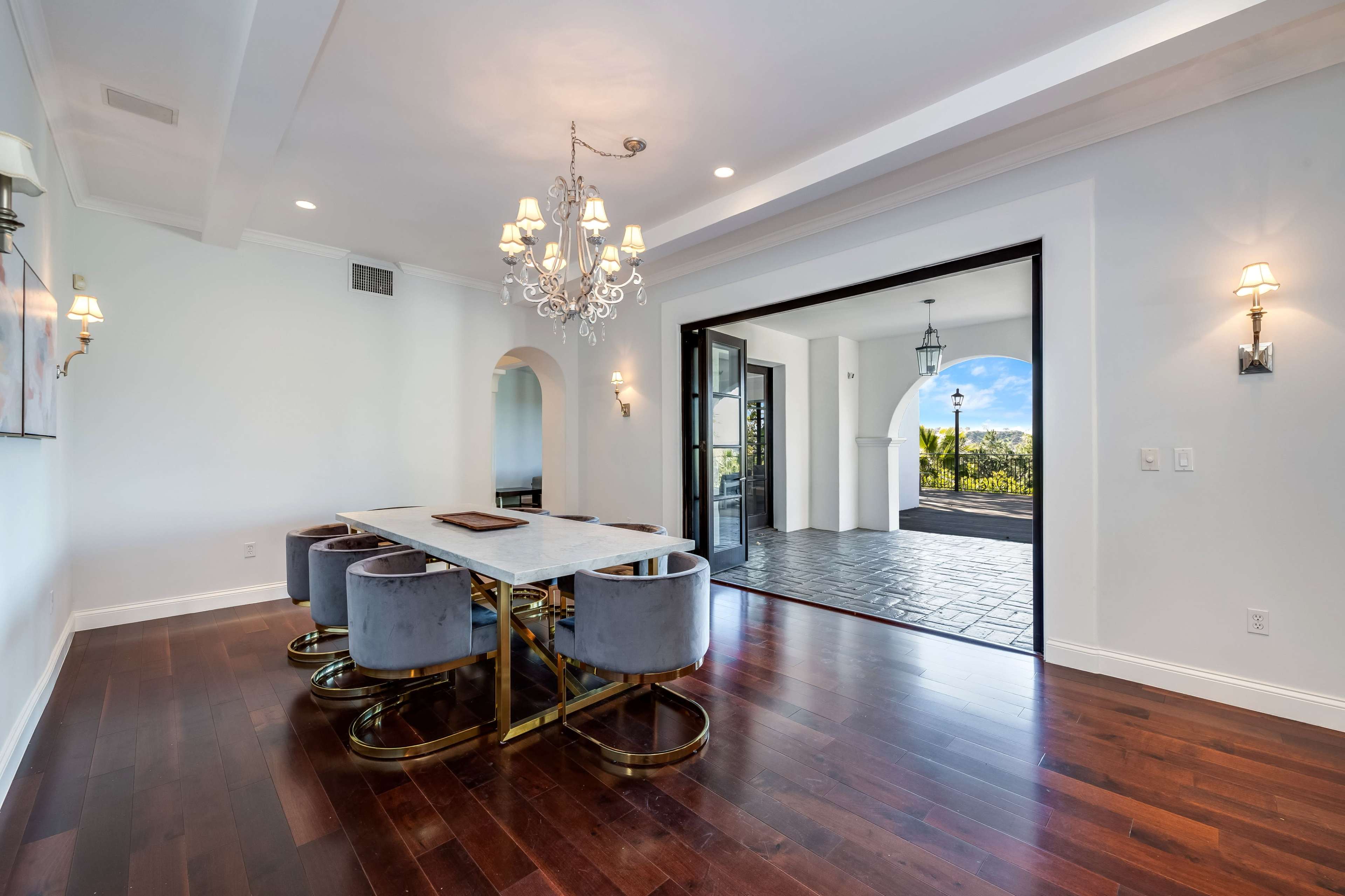 A dining area features a marble table with upholstered chairs and a chandelier, leading to an outdoor space through large glass doors.