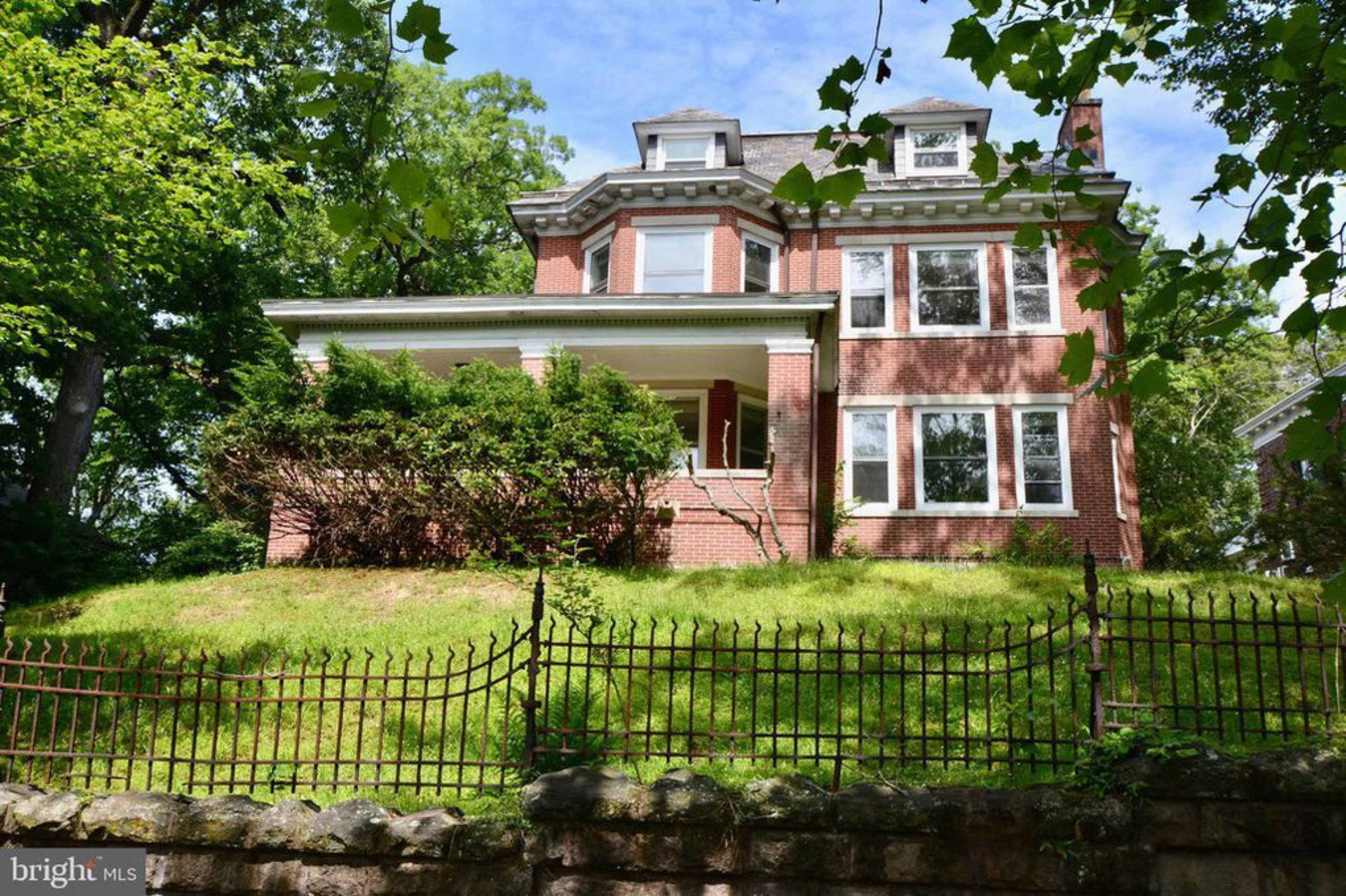 A brick house with multiple windows and a porch is situated on a grassy lot enclosed by an iron fence.