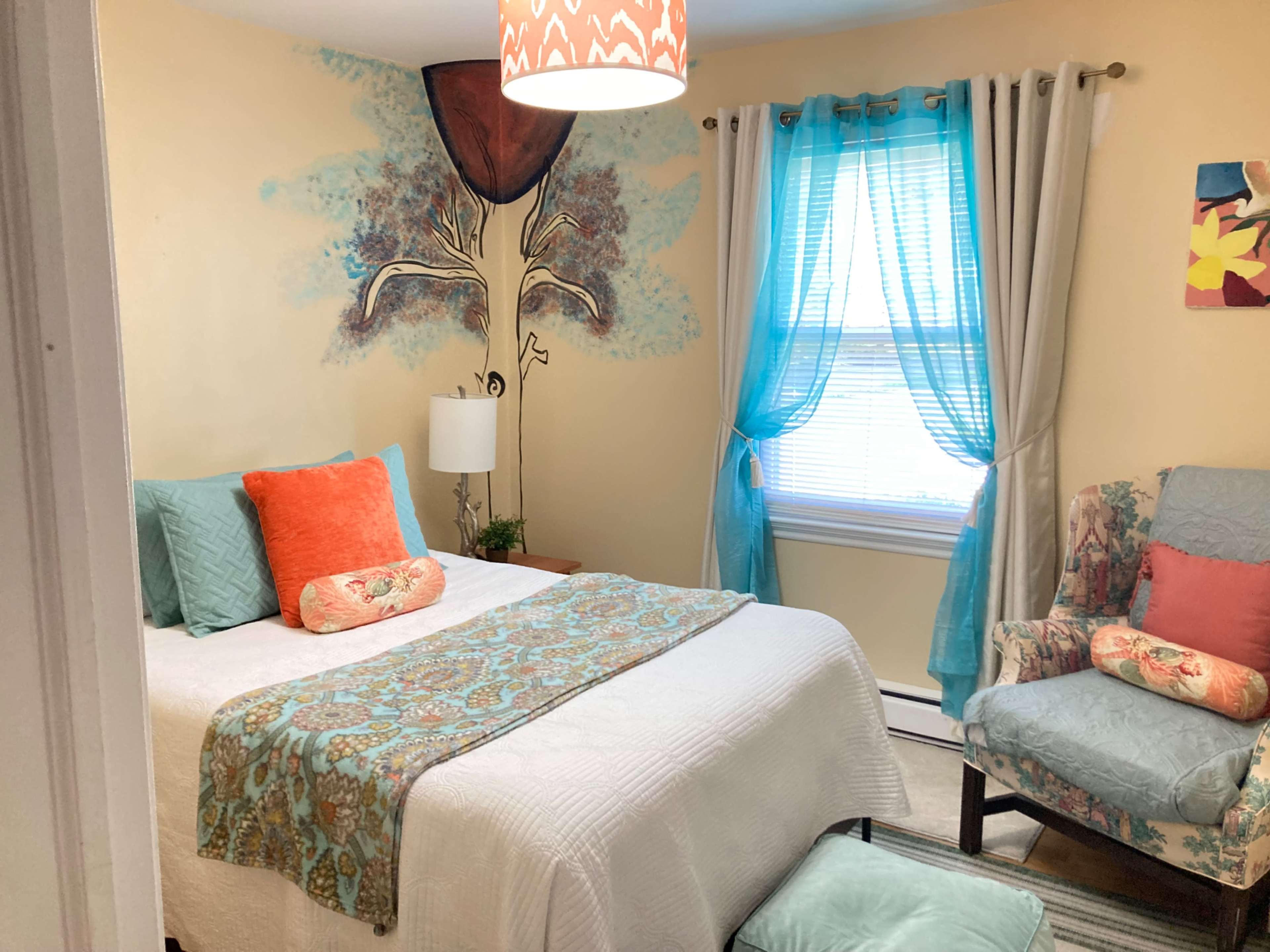 A small bedroom features a bed with colorful pillows, a decorative quilt, a chair with cushions, and a wall mural of a tree.
