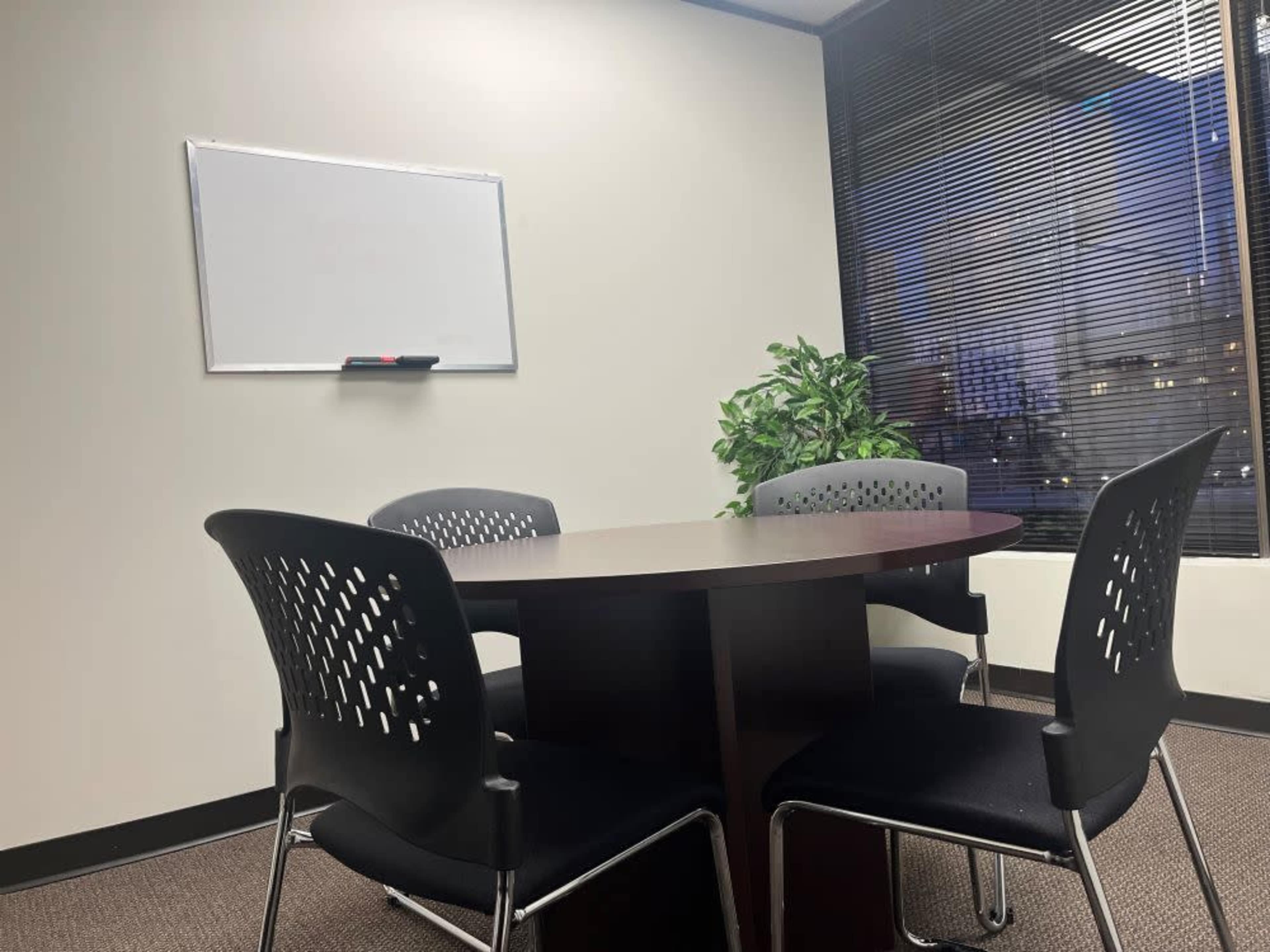 The image shows a small meeting room with a round table surrounded by four chairs, a whiteboard on the wall, and a potted plant near the window.