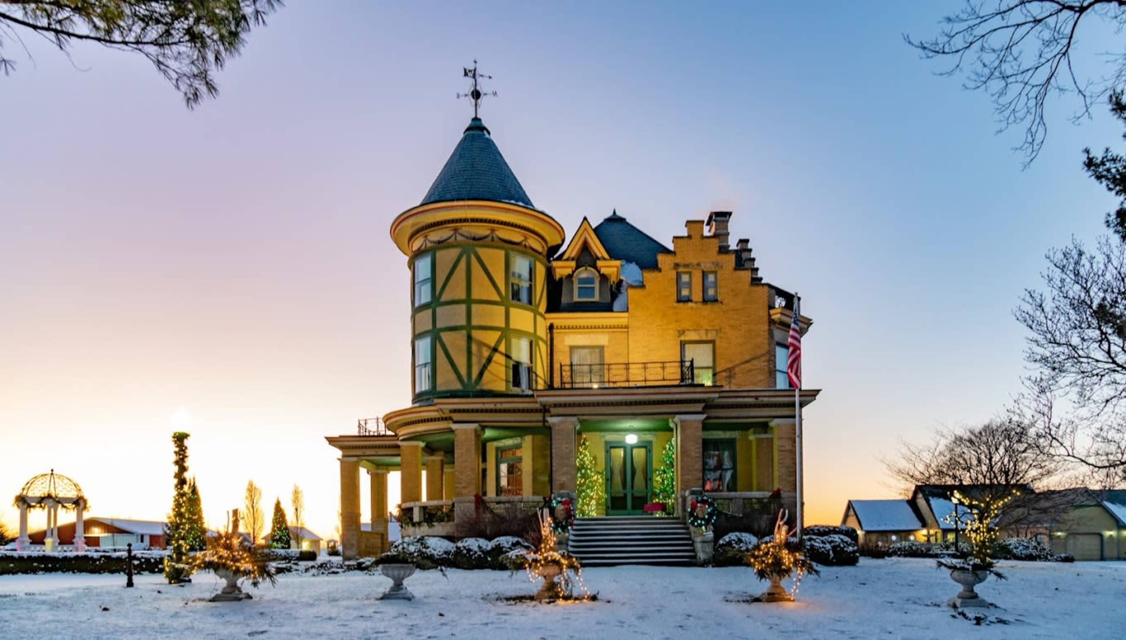 A large, ornate yellow mansion with a conical roof stands in a snowy landscape, adorned with festive lights and decorations.