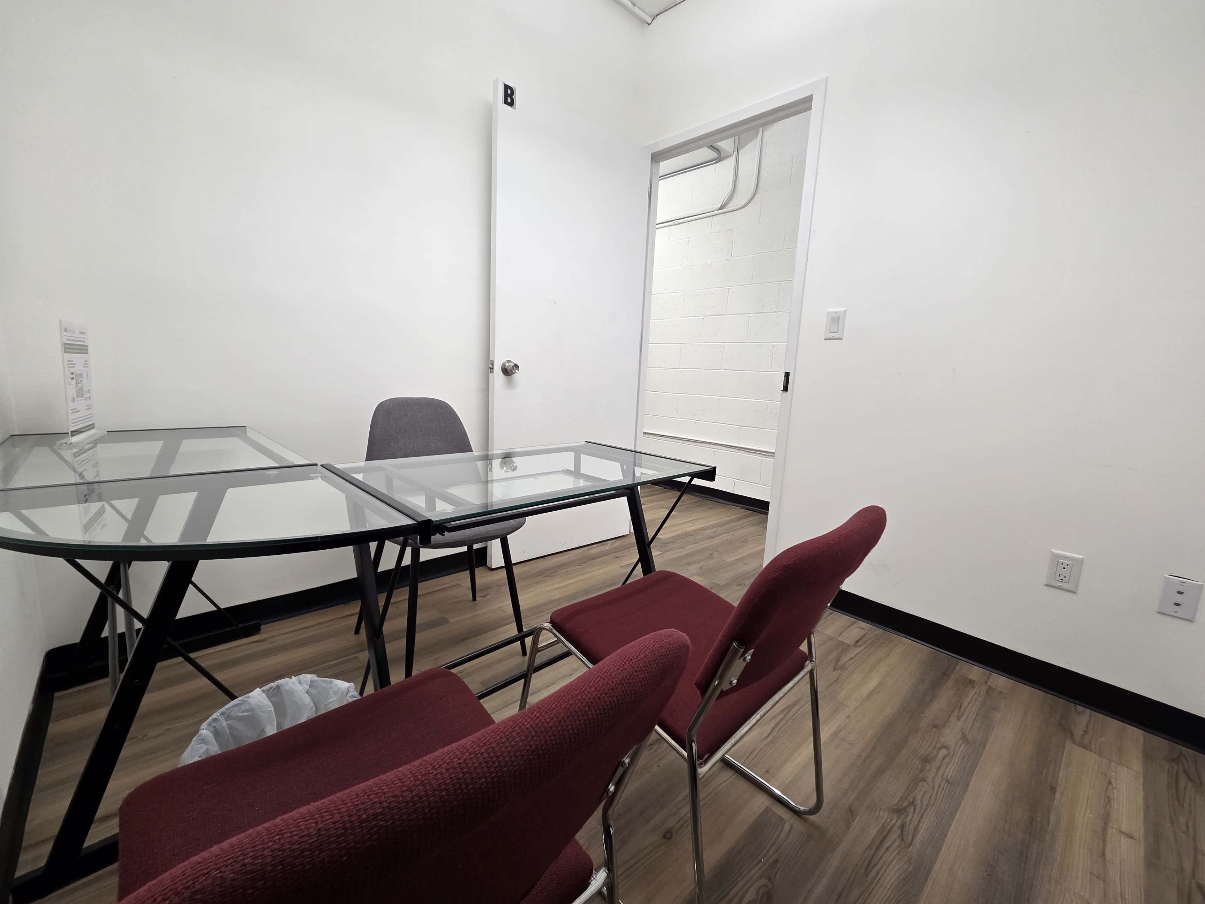 The image shows a small meeting room with two glass-top tables and several red chairs, facing an open door leading to a hallway.