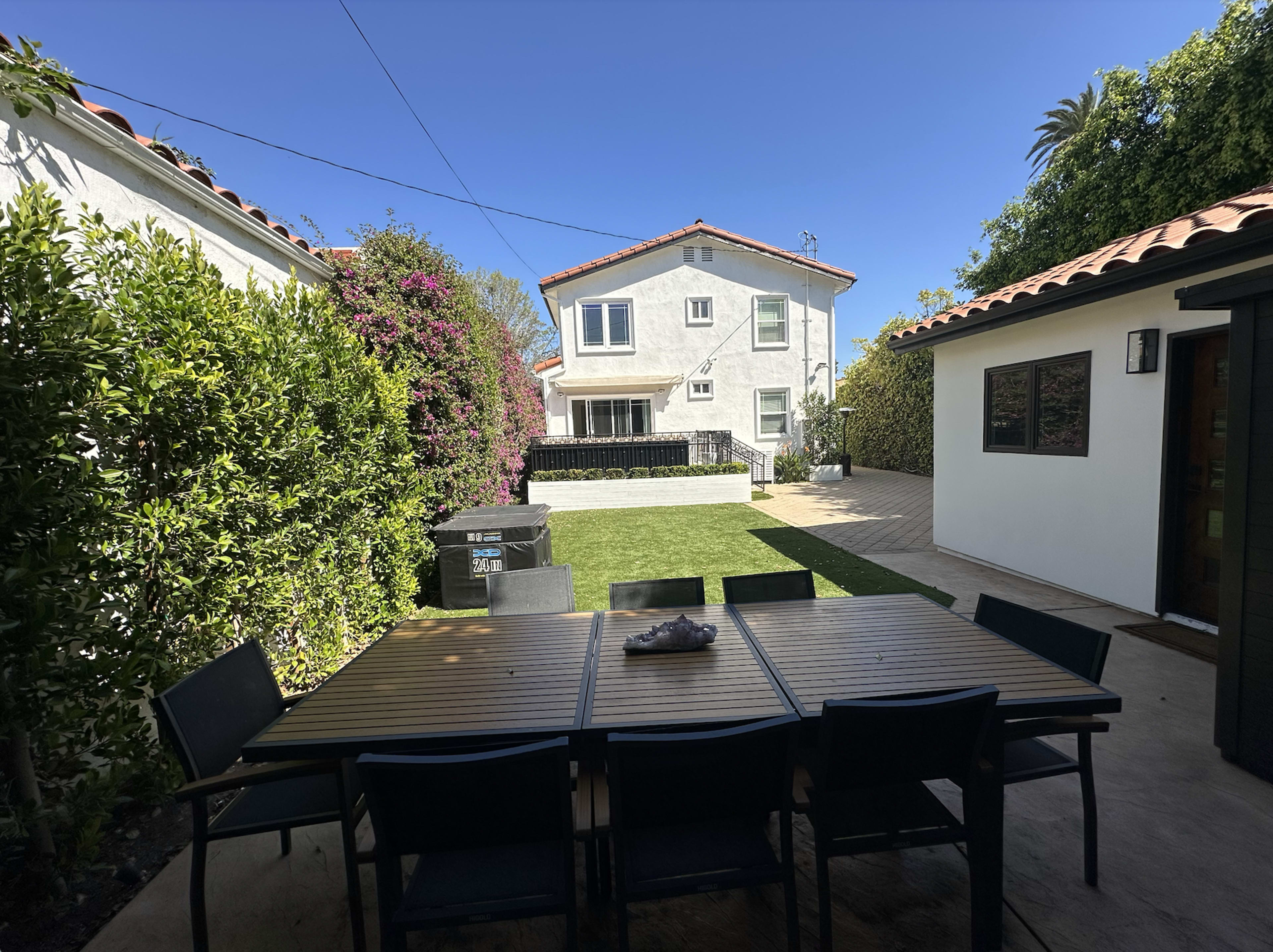 The image shows a backyard patio with a wooden dining table surrounded by black chairs, leading to a house with a garden and a lawn.