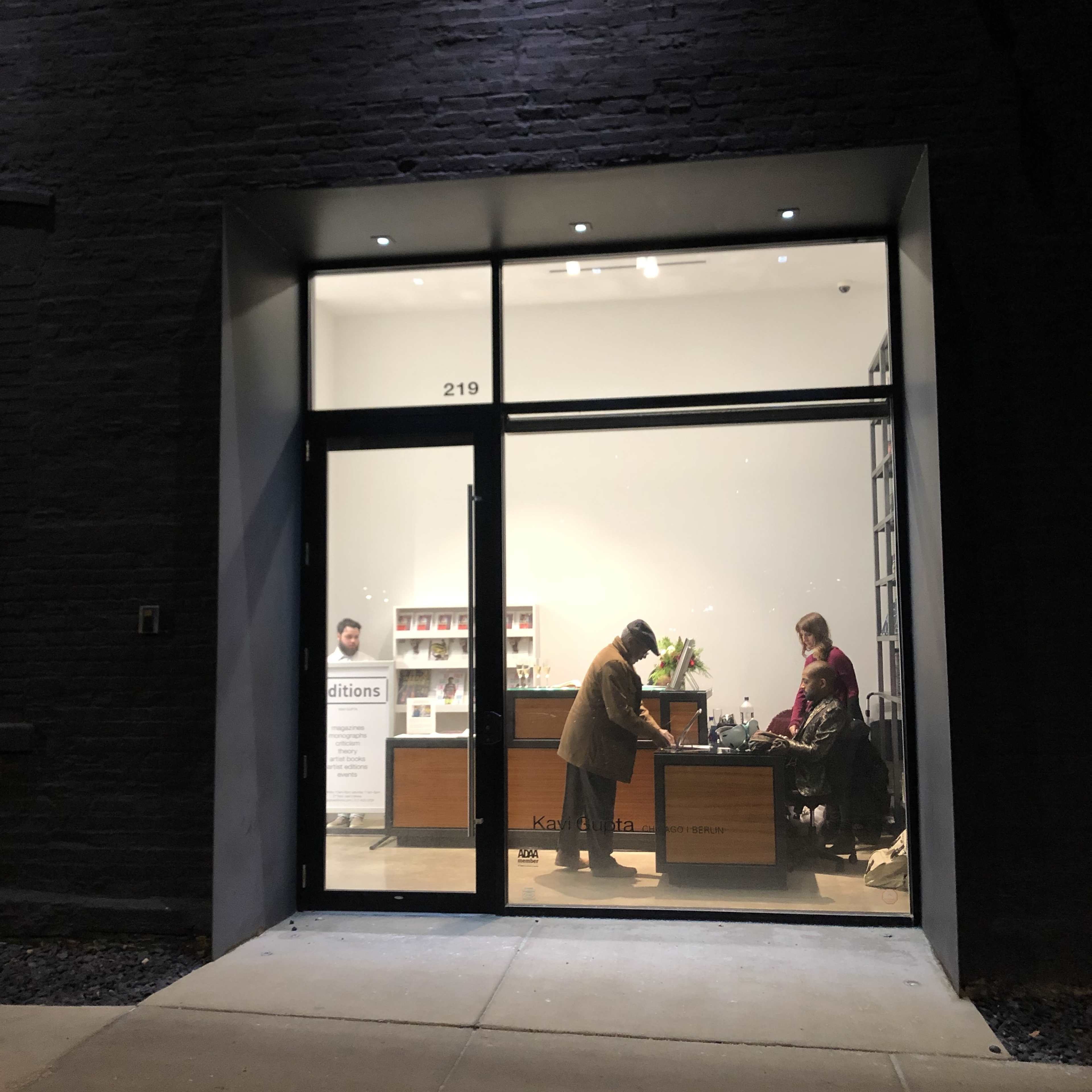 The image shows a cozy reception area with a large window, displaying three people engaged in conversation and work at a front desk.
