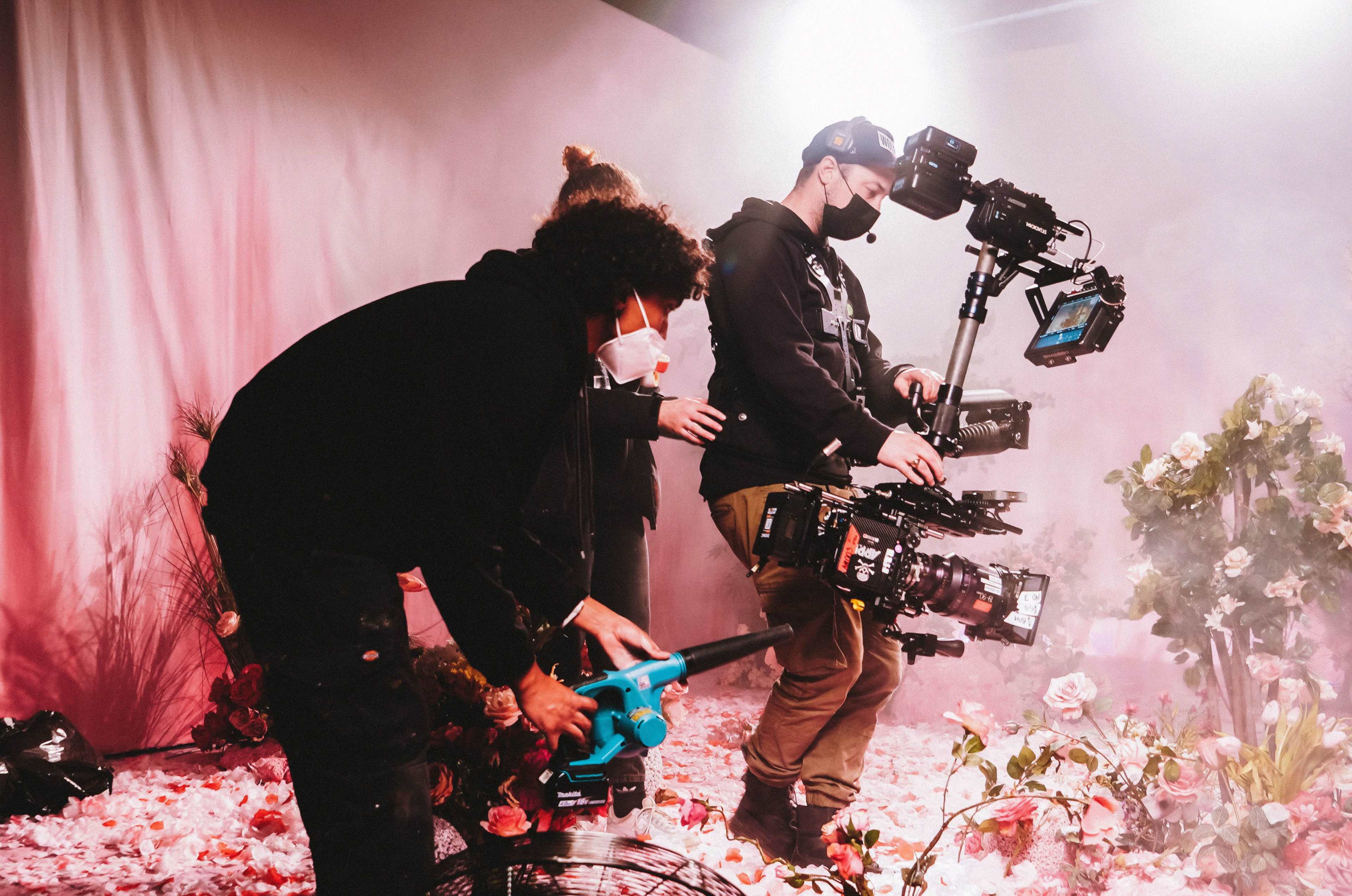 Two filmmakers operate camera equipment in a studio surrounded by flowers and mist, while a third person uses a leaf blower.