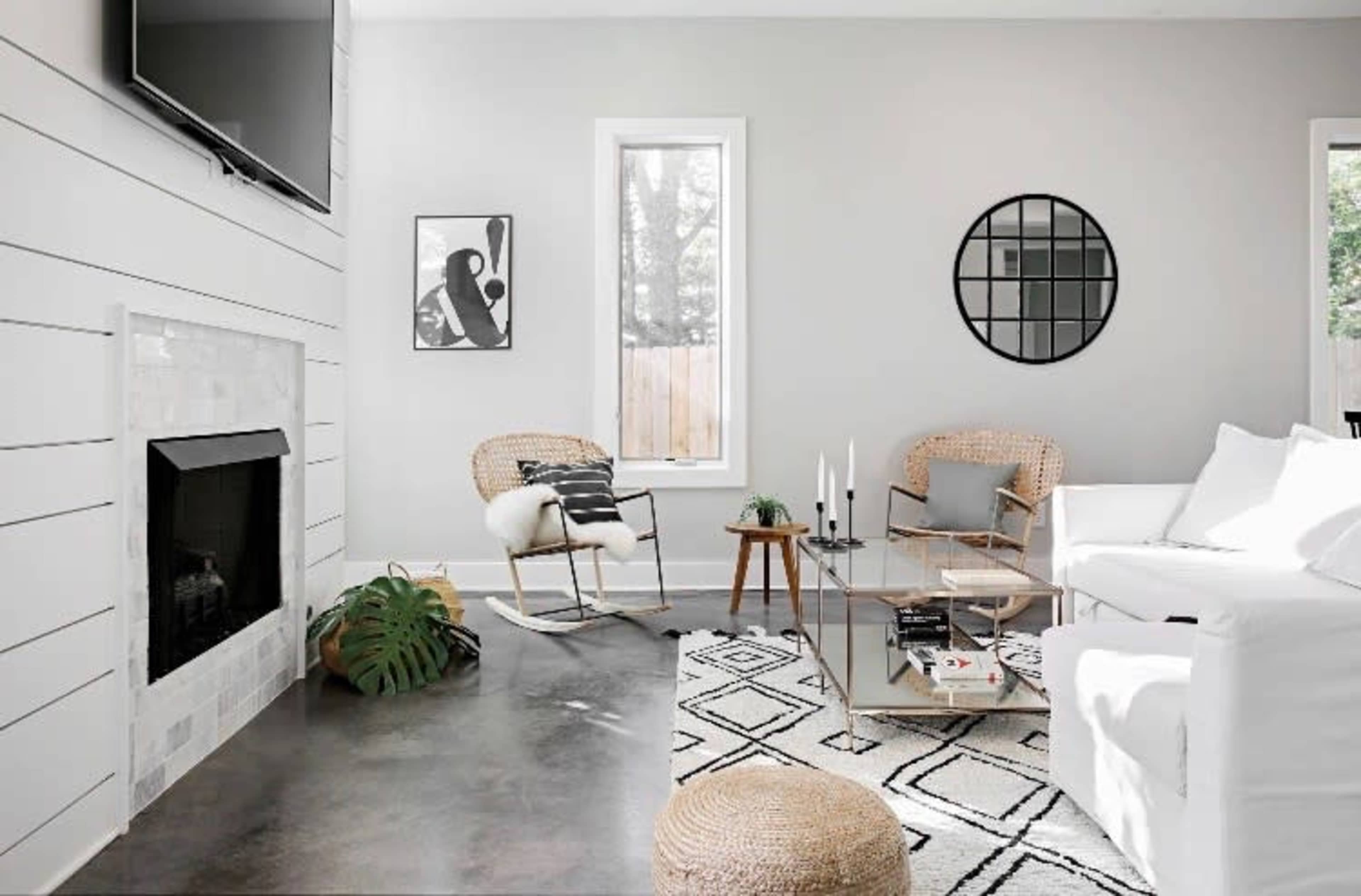 A modern living room featuring a white sofa, a glass coffee table, a fireplace, and a large window, all set against light gray walls.