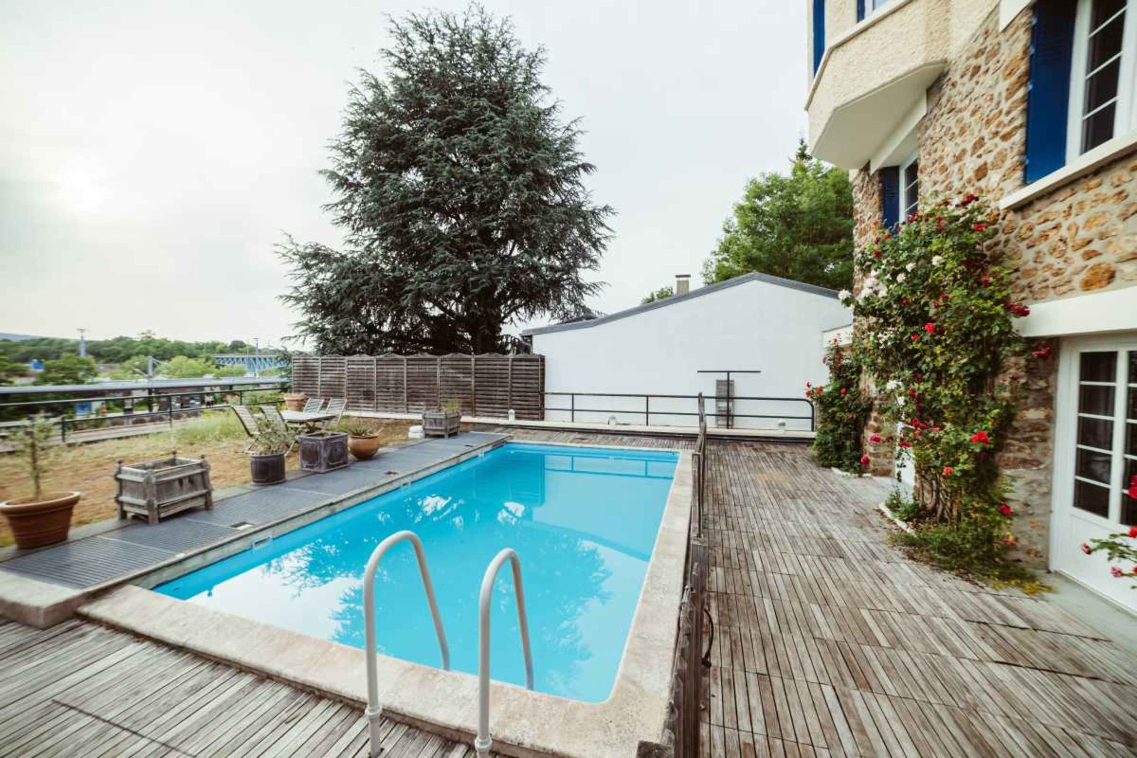 The image shows a rectangular swimming pool surrounded by a wooden deck, adjacent to a stone building and landscaped with potted plants.