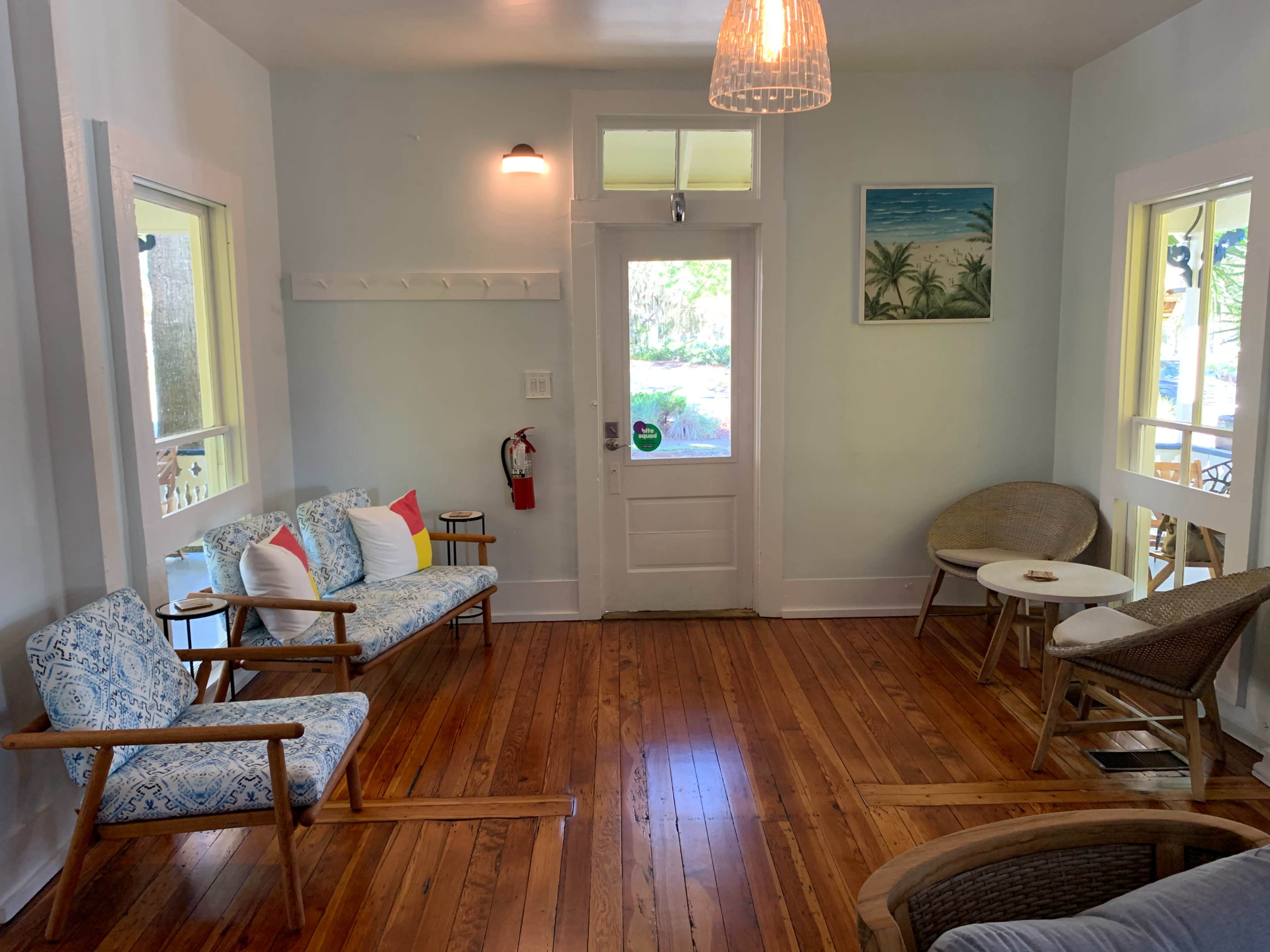 The image shows a cozy sitting area with wooden floors, a door leading outside, and several seating options, including a sofa and chairs, along with a small table.