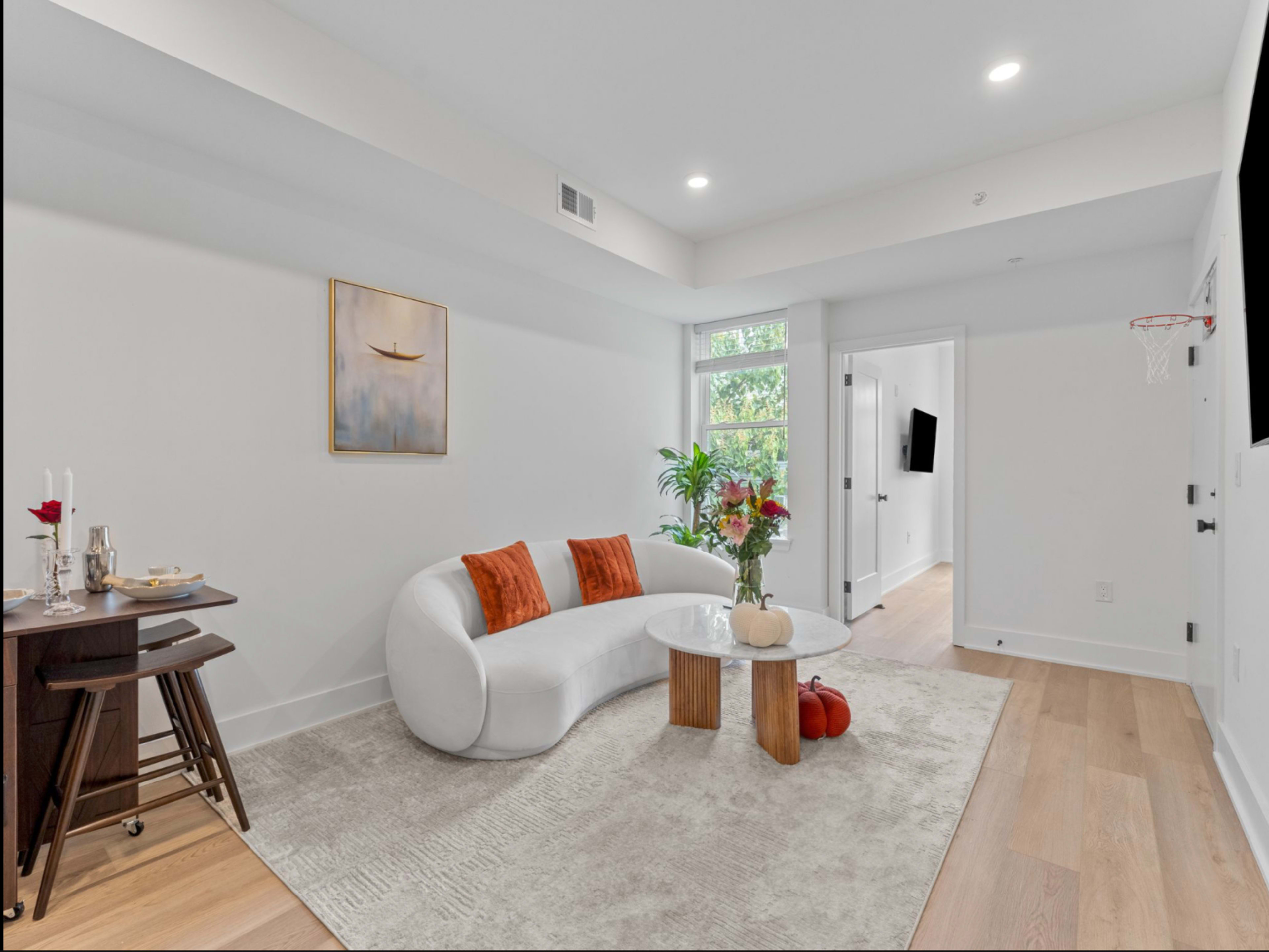The image shows a modern living room with a curved white sofa, a glass coffee table, and a small dining area with a set table.