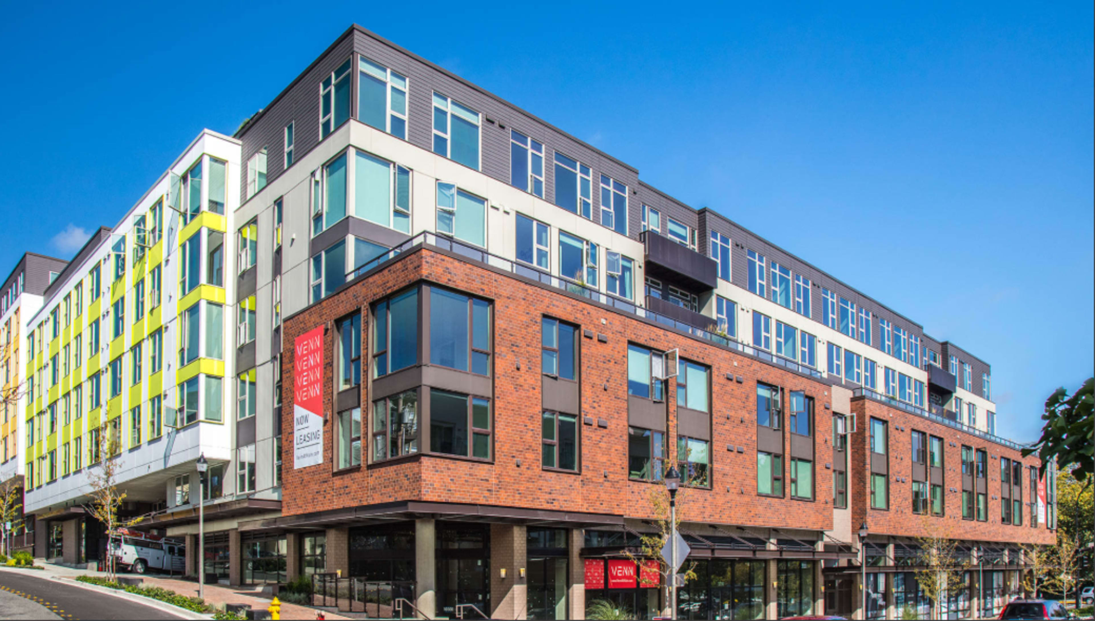 The image shows a modern multi-story building with a combination of brick and glass facades, featuring commercial spaces on the ground floor and residential units above.