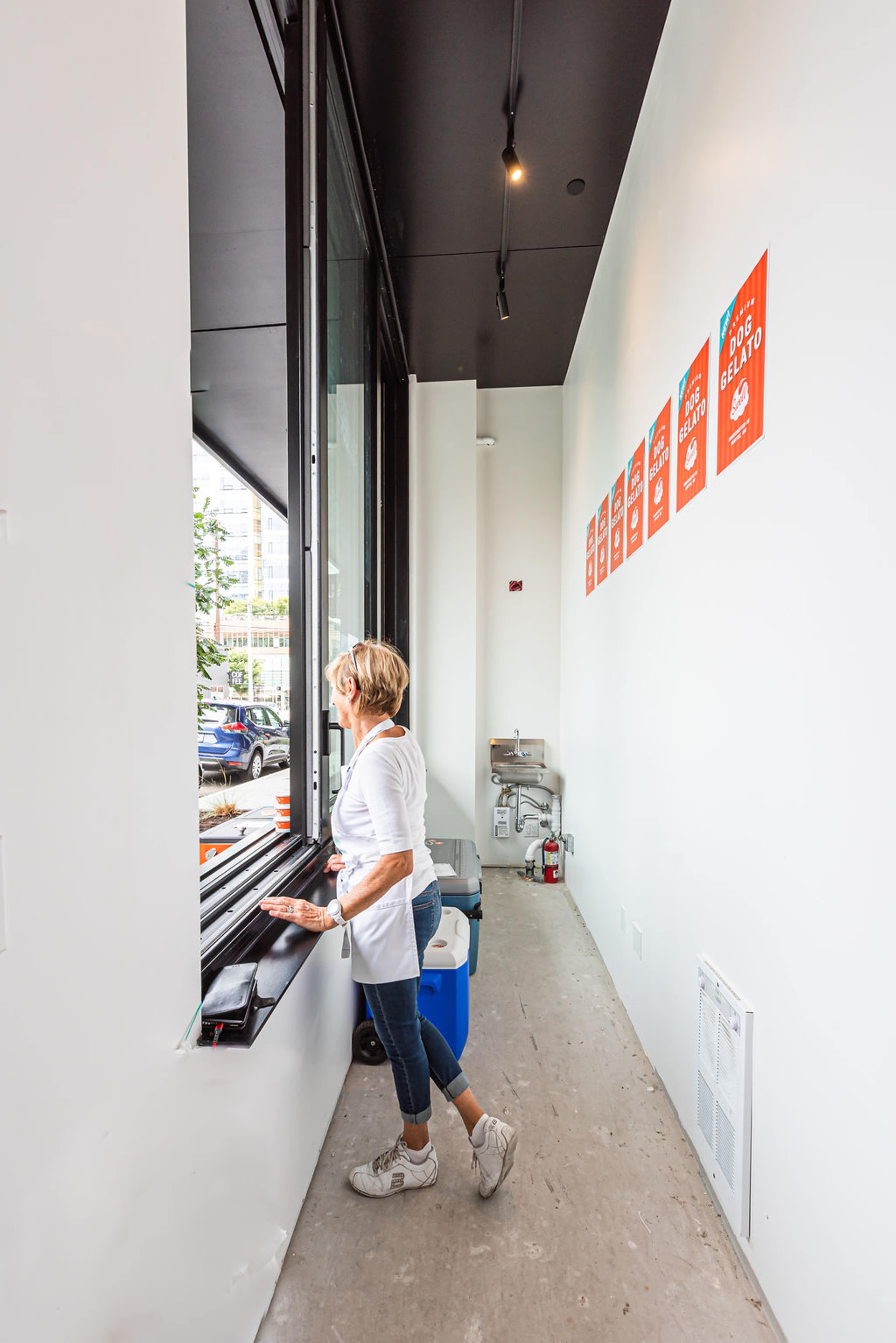 A woman stands by a large window in a minimalist room, looking out onto a street while several colorful posters hang on the wall.