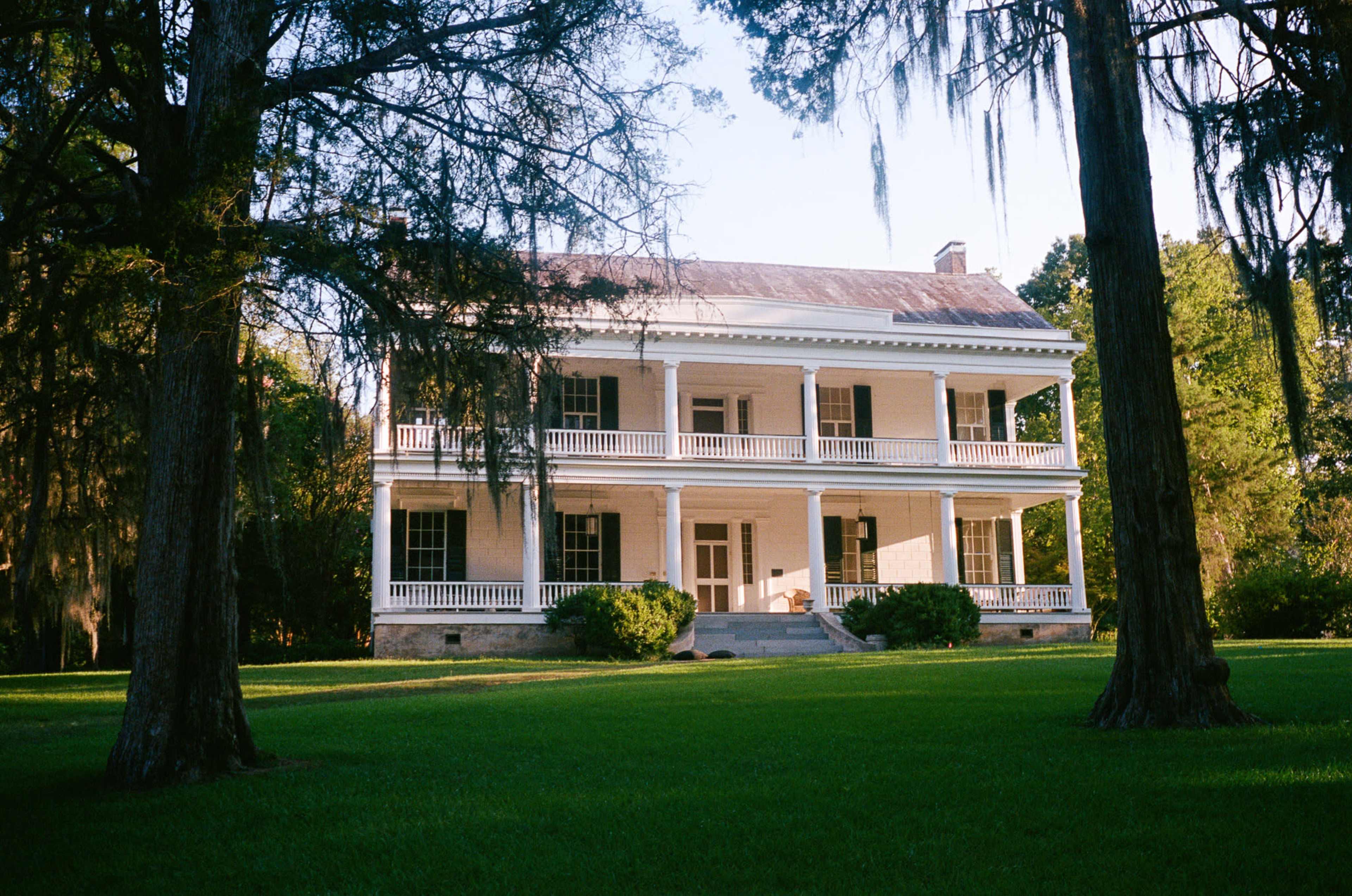 A two-story white plantation house with a large front porch is surrounded by green grass and tall trees.