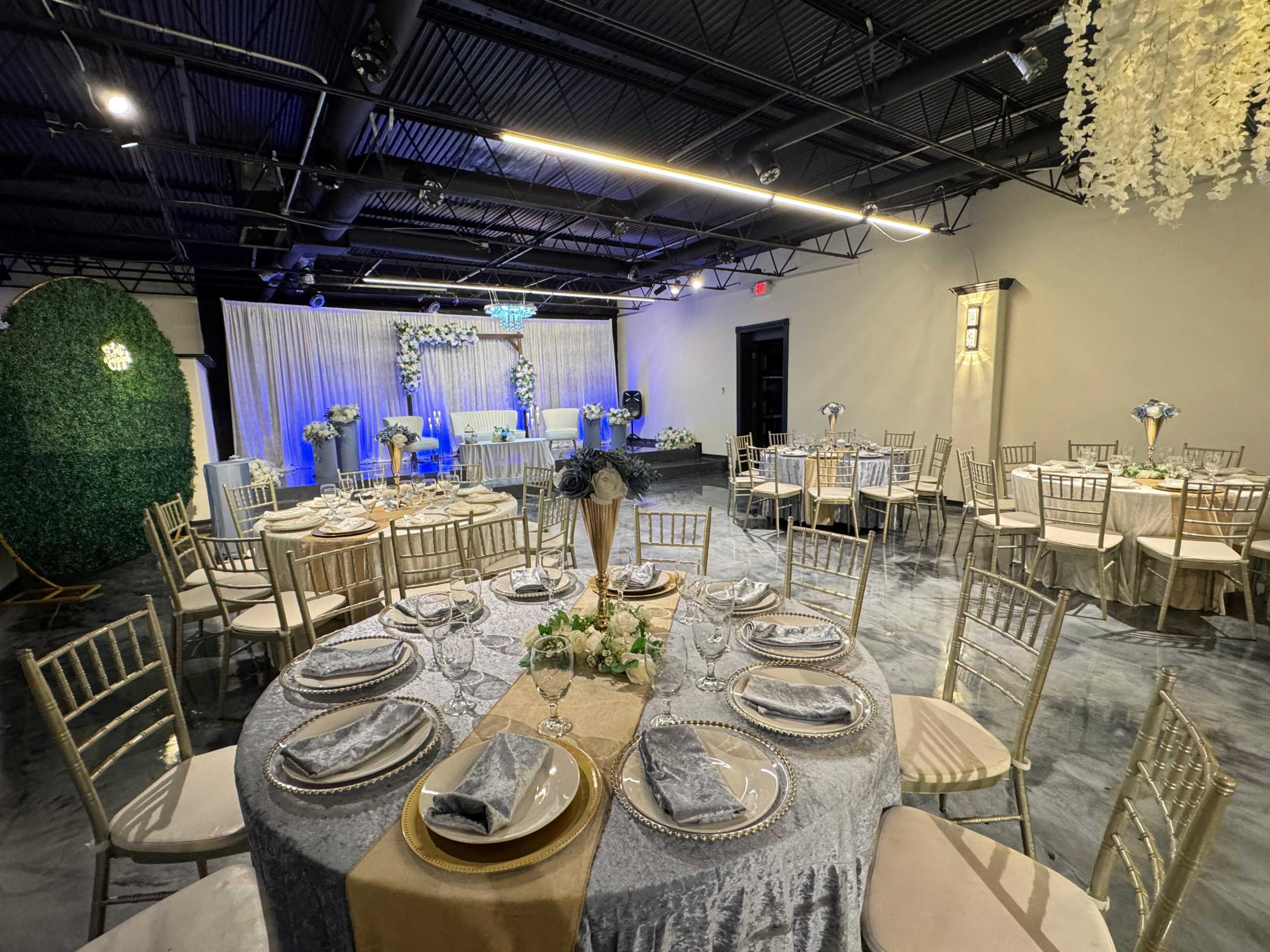 The image shows a decorated event space with elegantly set round tables, each featuring a tablecloth, plates, and napkins, along with a staging area adorned with floral arrangements and ambient lighting.