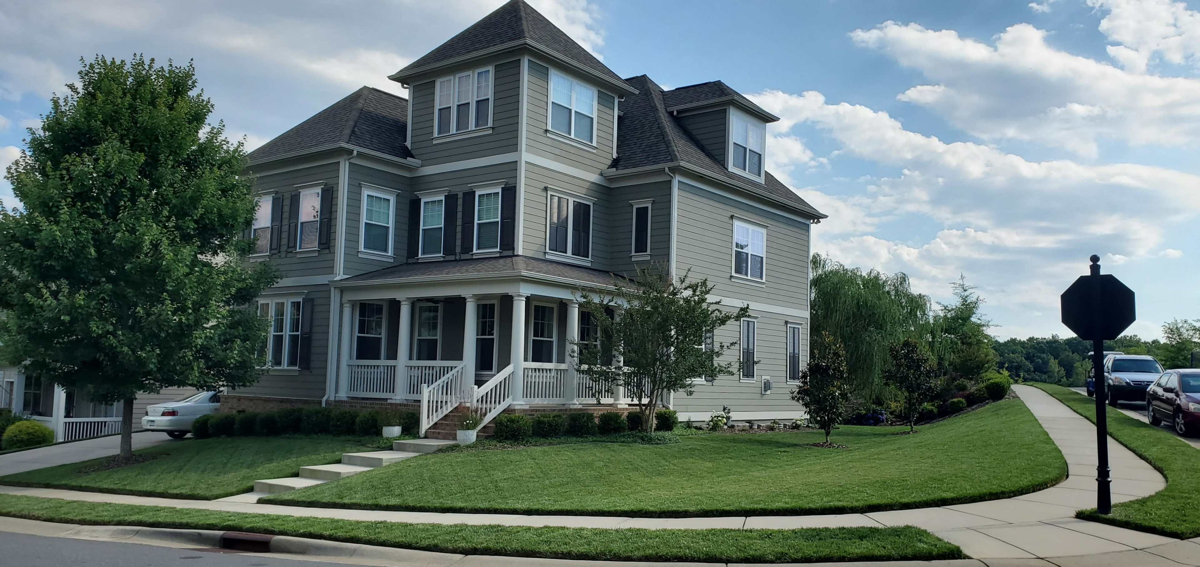 A large, two-story house with a wraparound porch, surrounded by manicured grass and decorative shrubs, stands at the corner of a street.