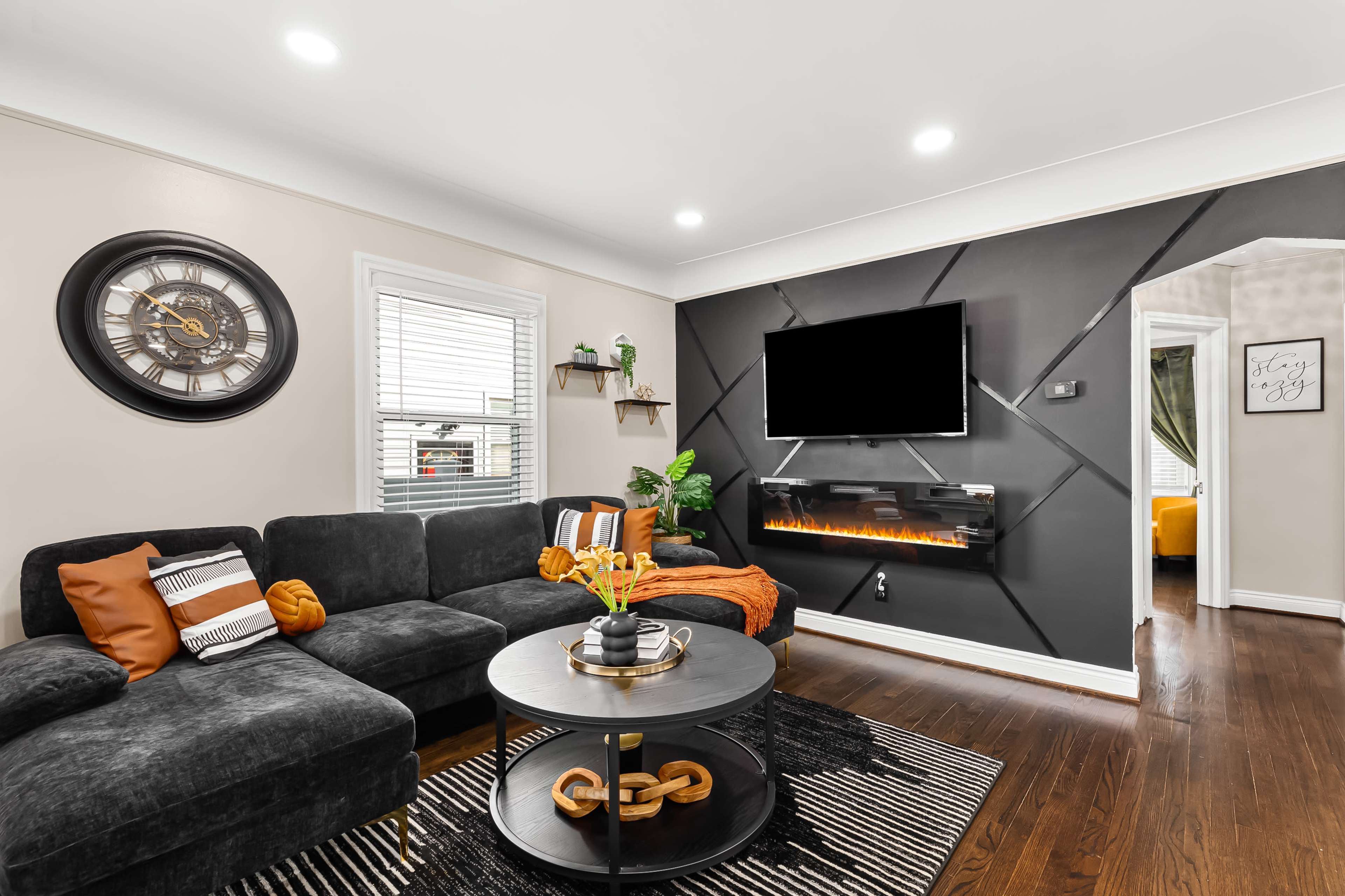 The living room features a black sectional sofa, a round coffee table with a decorative chain, and an accent wall with a mounted TV and a modern electric fireplace.