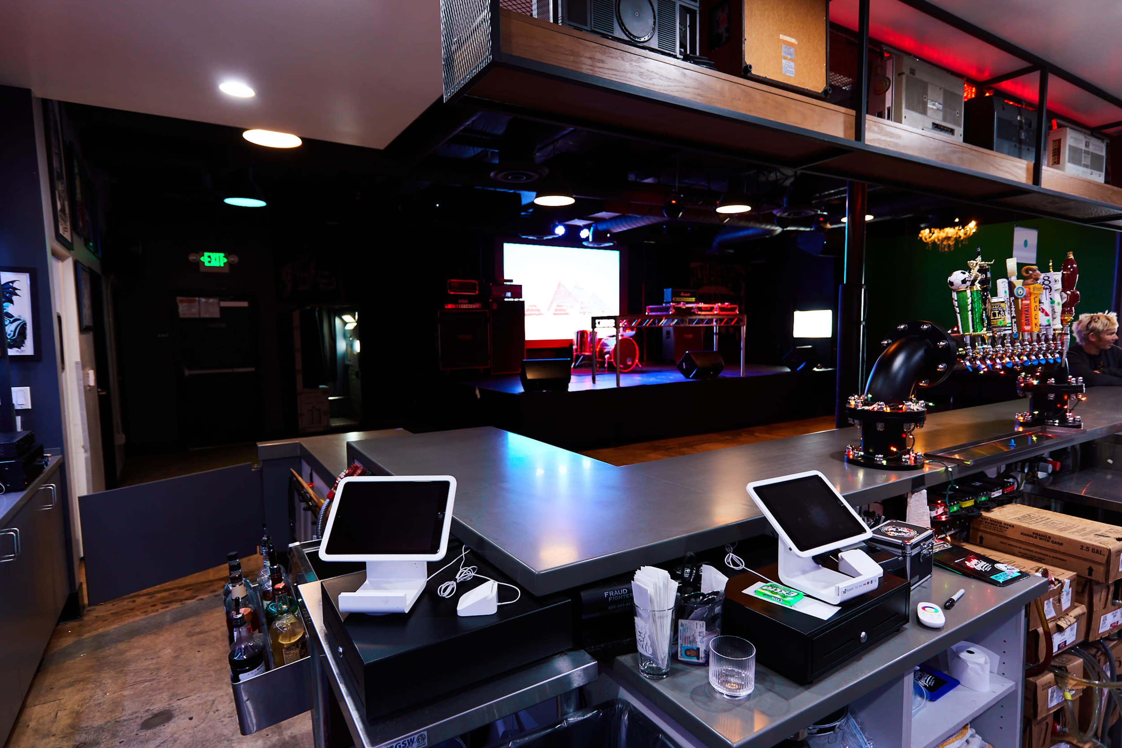 A bar area with digital point-of-sale terminals in the foreground, leading to a performance stage in the background.