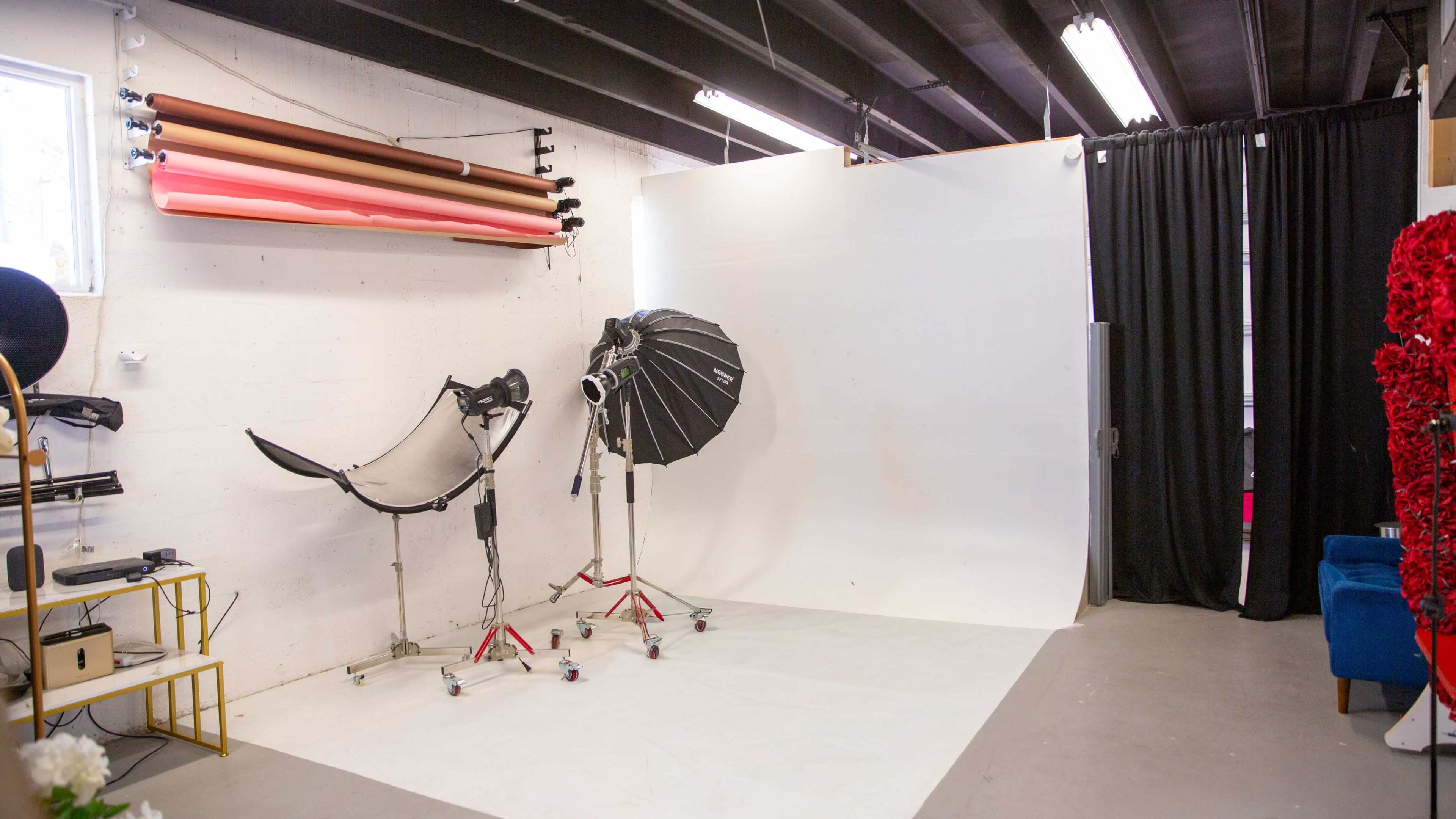 The image shows a photography studio featuring a white backdrop, lighting equipment on stands, and colorful backdrop rolls mounted on the wall.