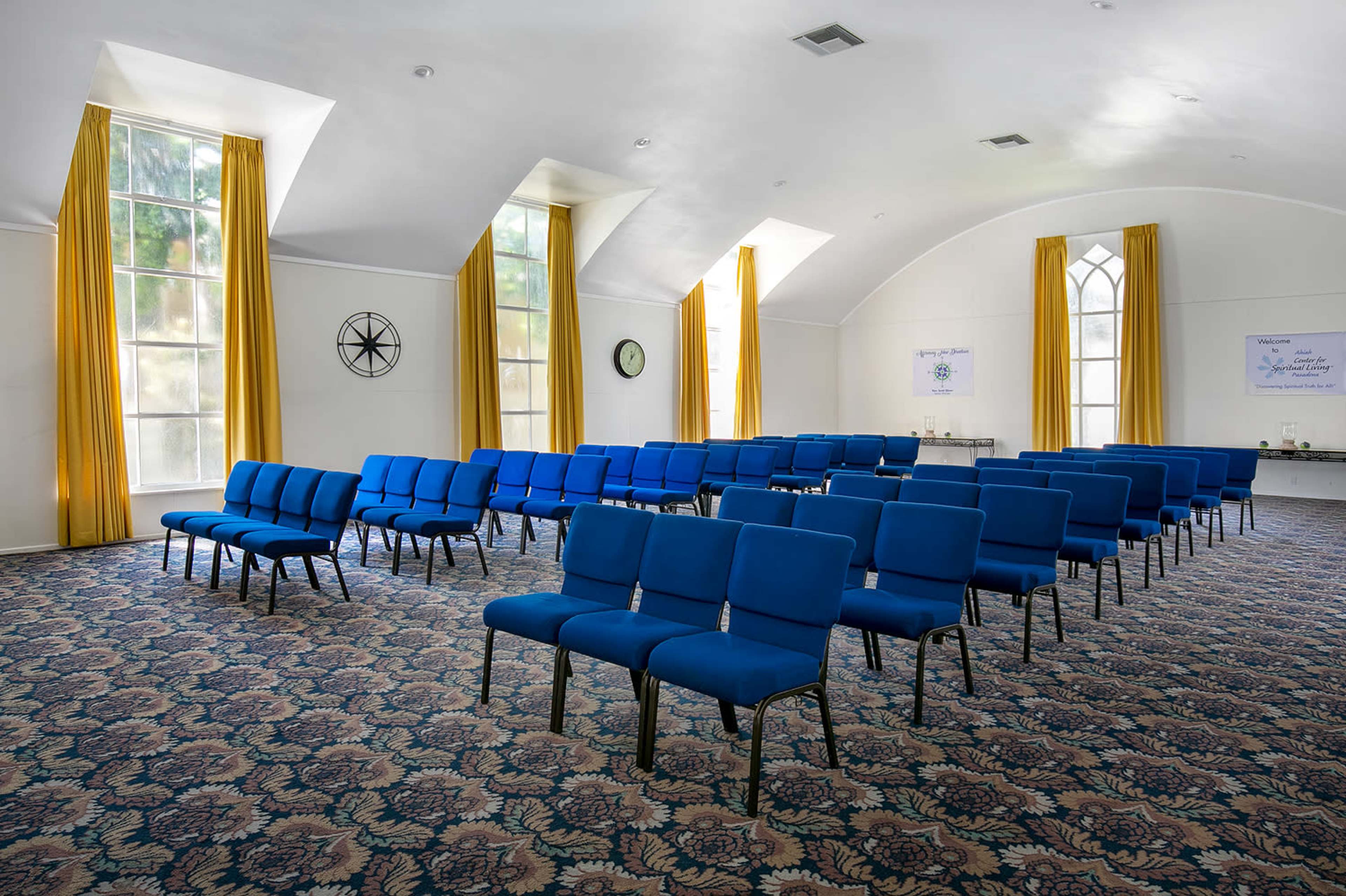 The image shows a room with blue chairs arranged in rows, large windows with yellow curtains, and a patterned carpet.