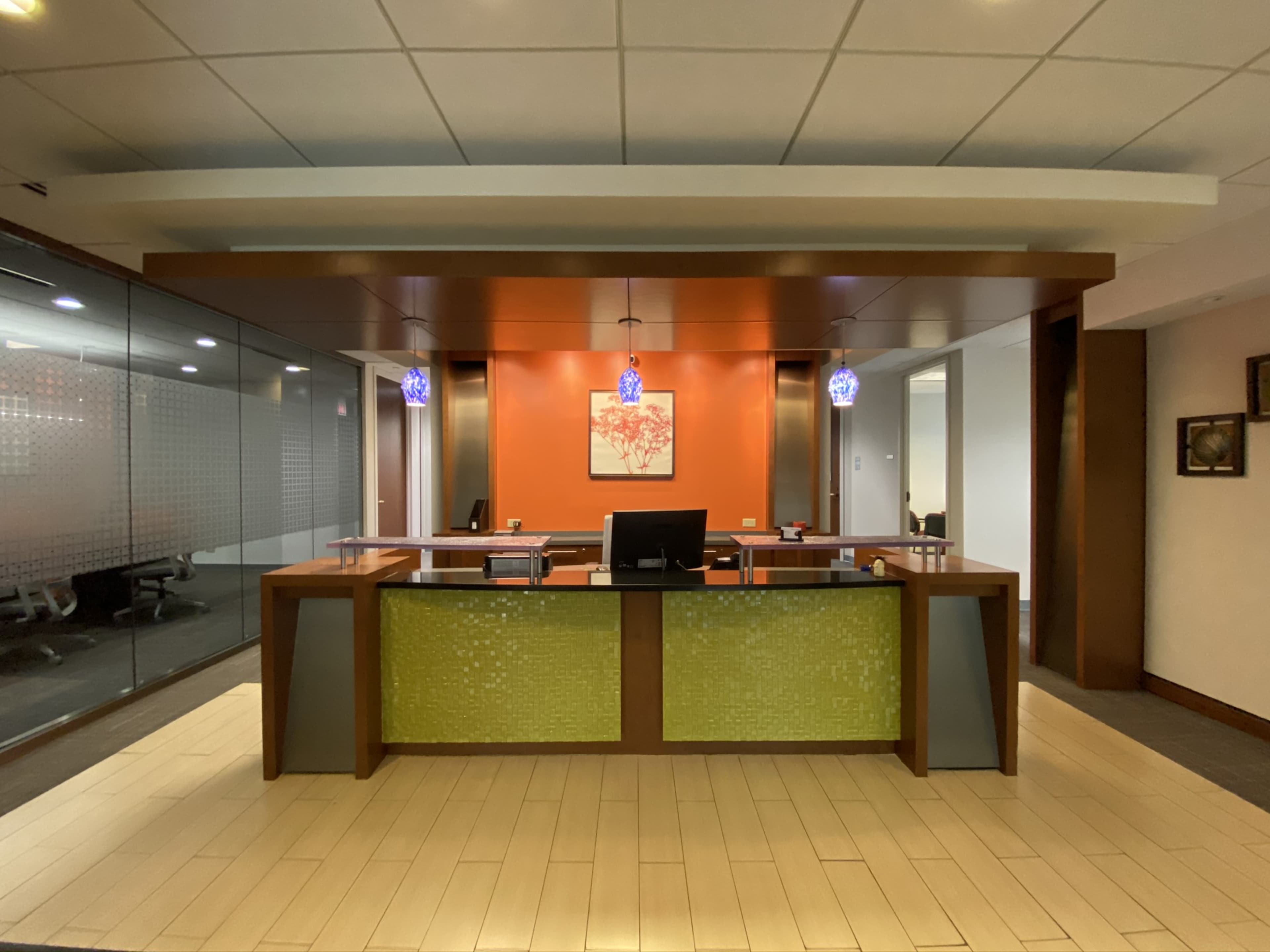 The image shows a modern office reception area with a wooden front desk, glass partitions, and decorative lighting.