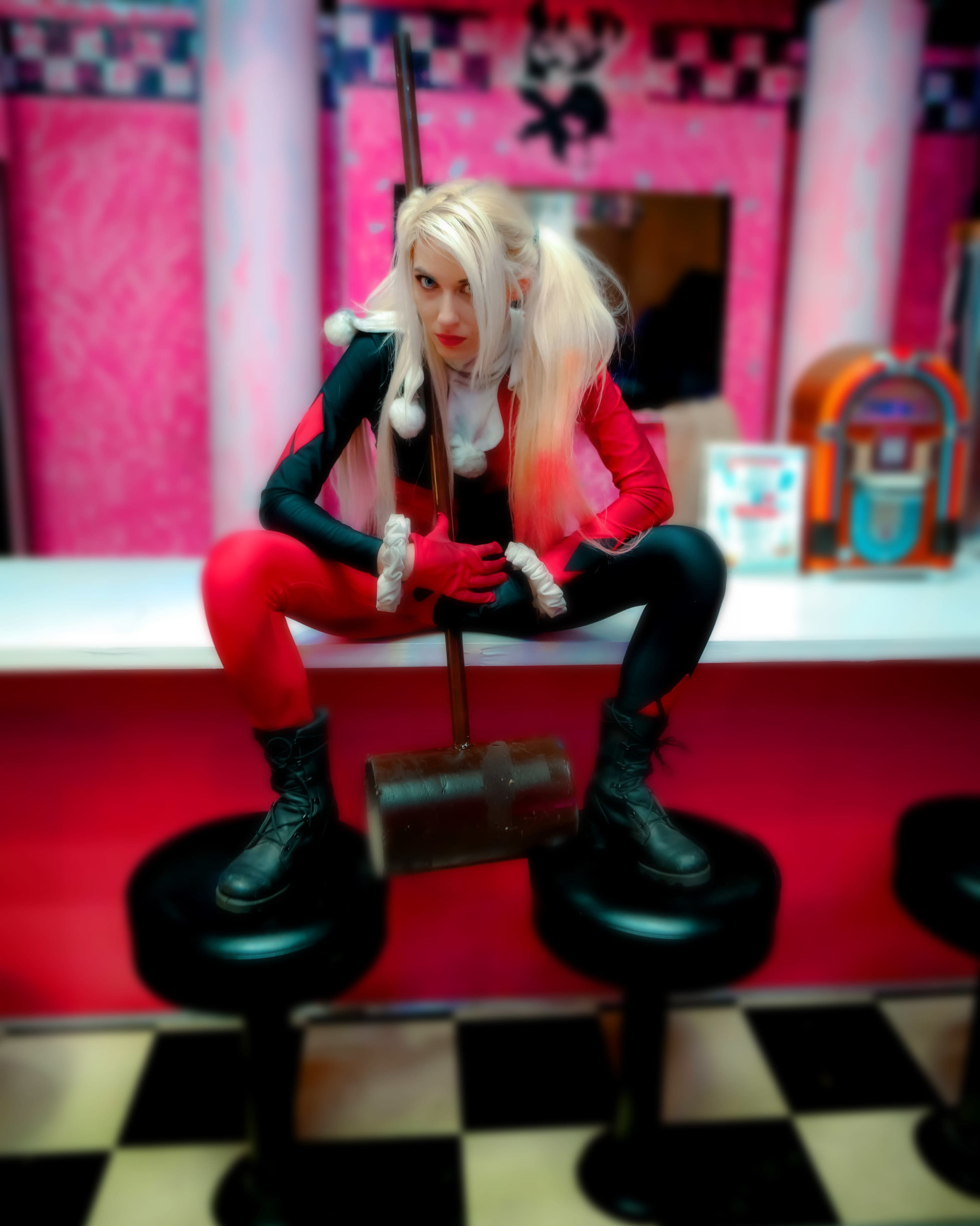 A person dressed as a character with a red and black outfit poses on a bar stool while holding a large mallet in a retro diner setting.