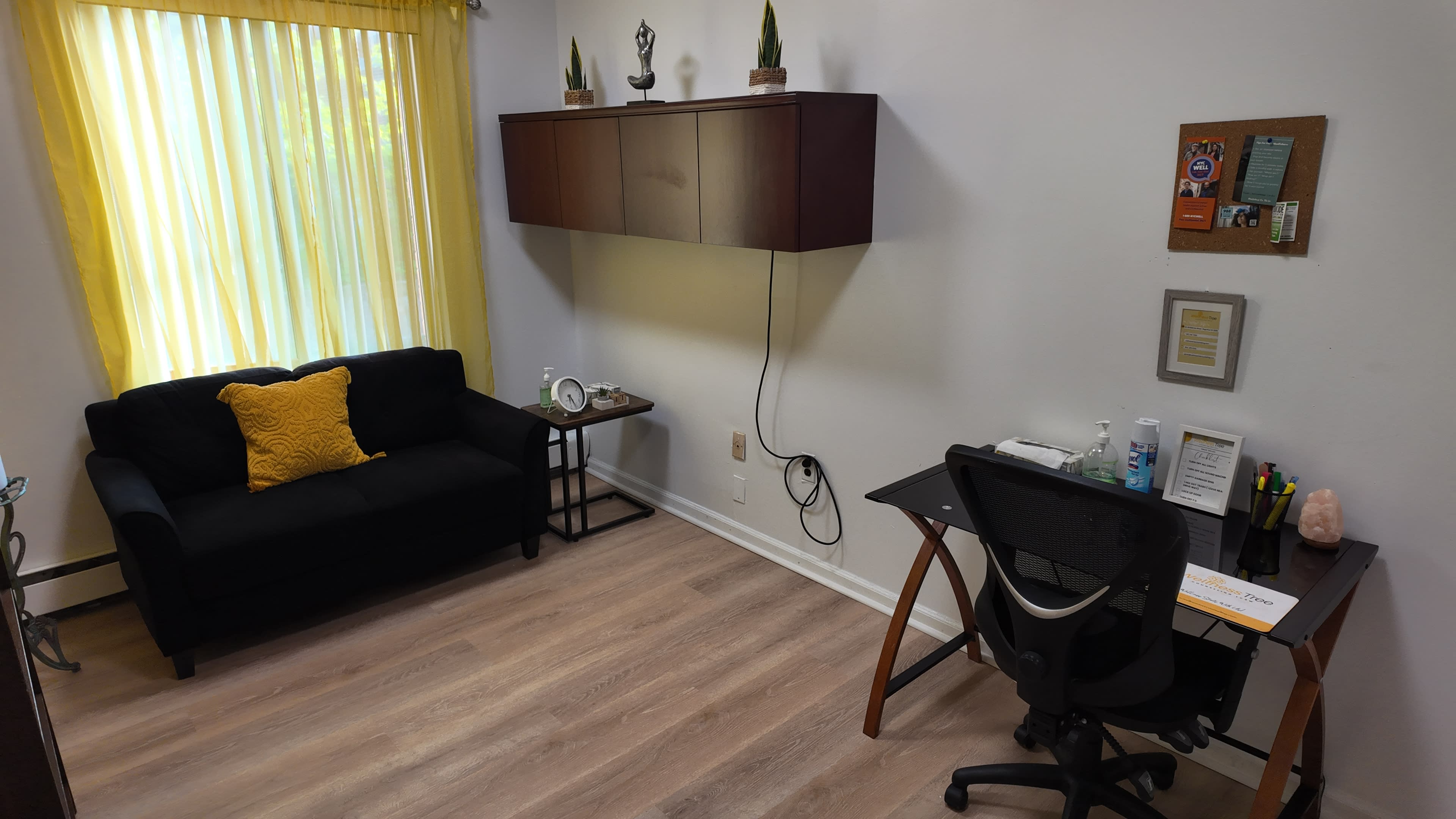The image shows a small office space with a black couch, a yellow decorative pillow, a desk equipped with stationery, and a wall-mounted cabinet.