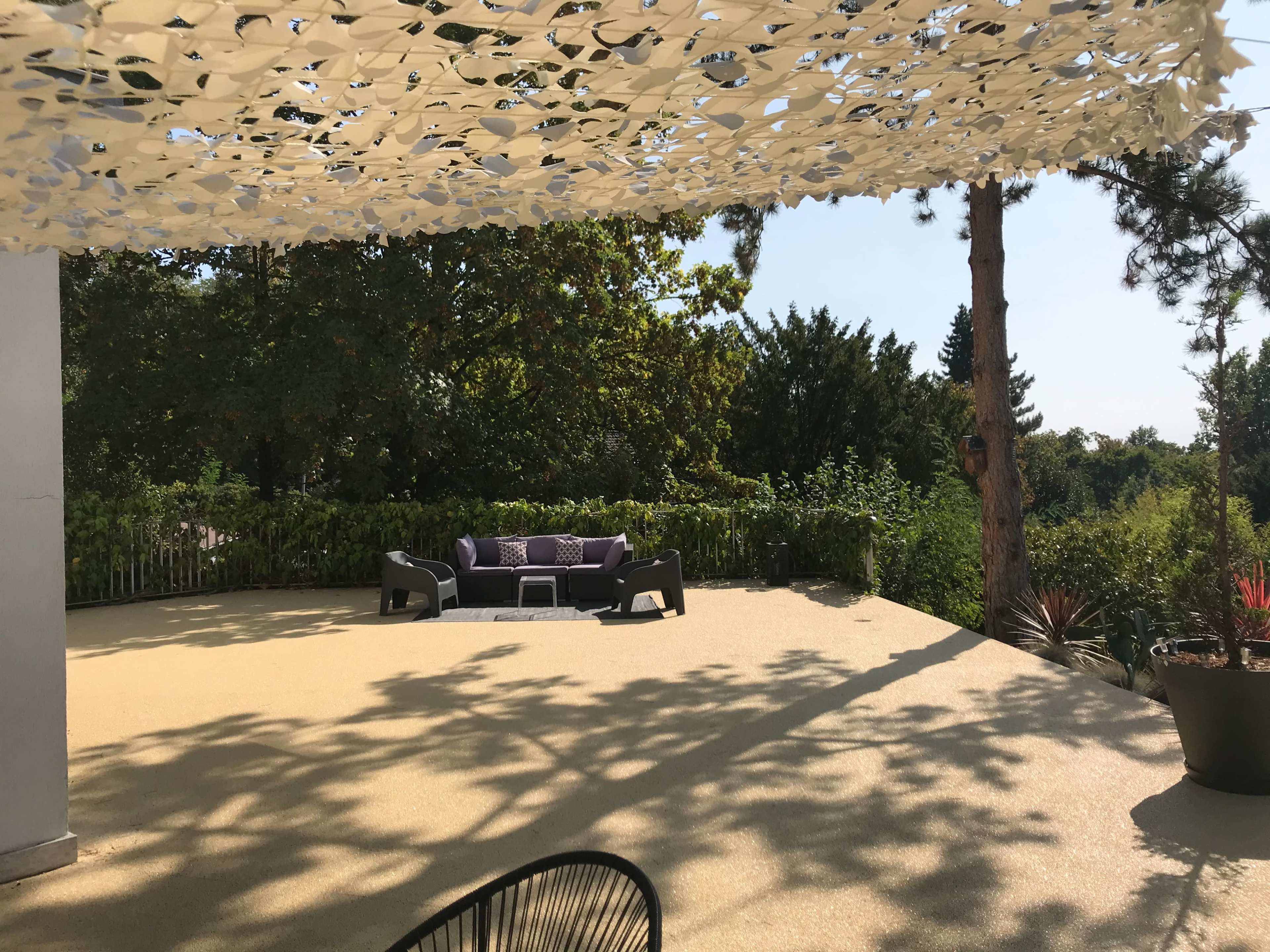 A shaded outdoor patio features a seating area with dark furniture surrounded by greenery.