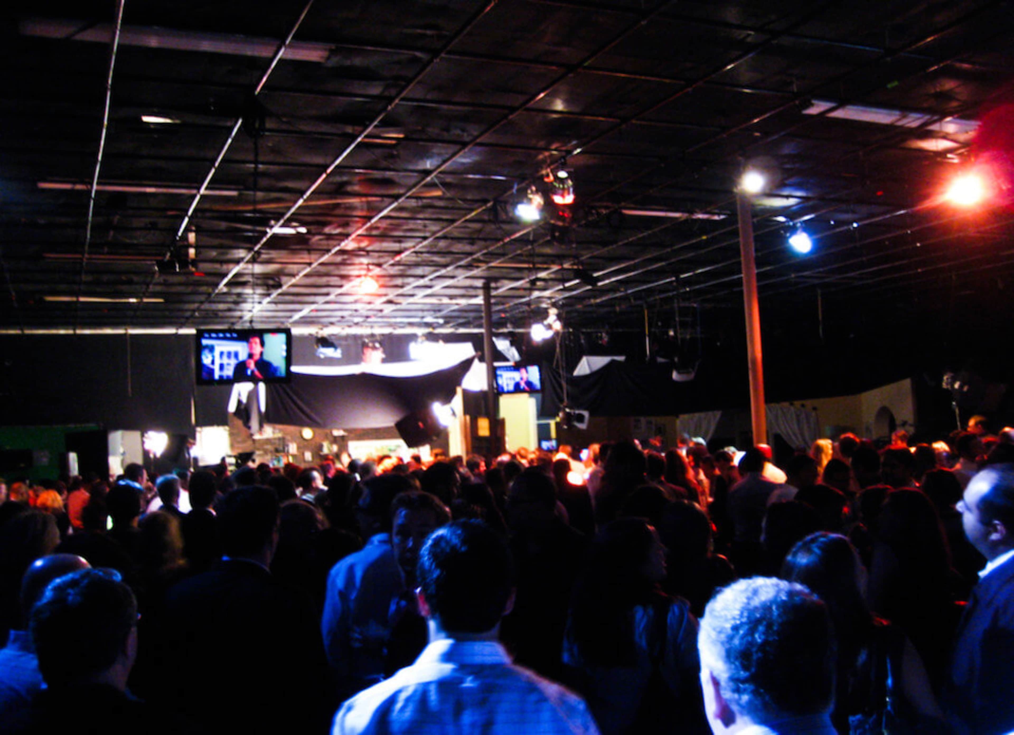 A crowded indoor space filled with people, featuring dim lighting and multiple screens displaying images.