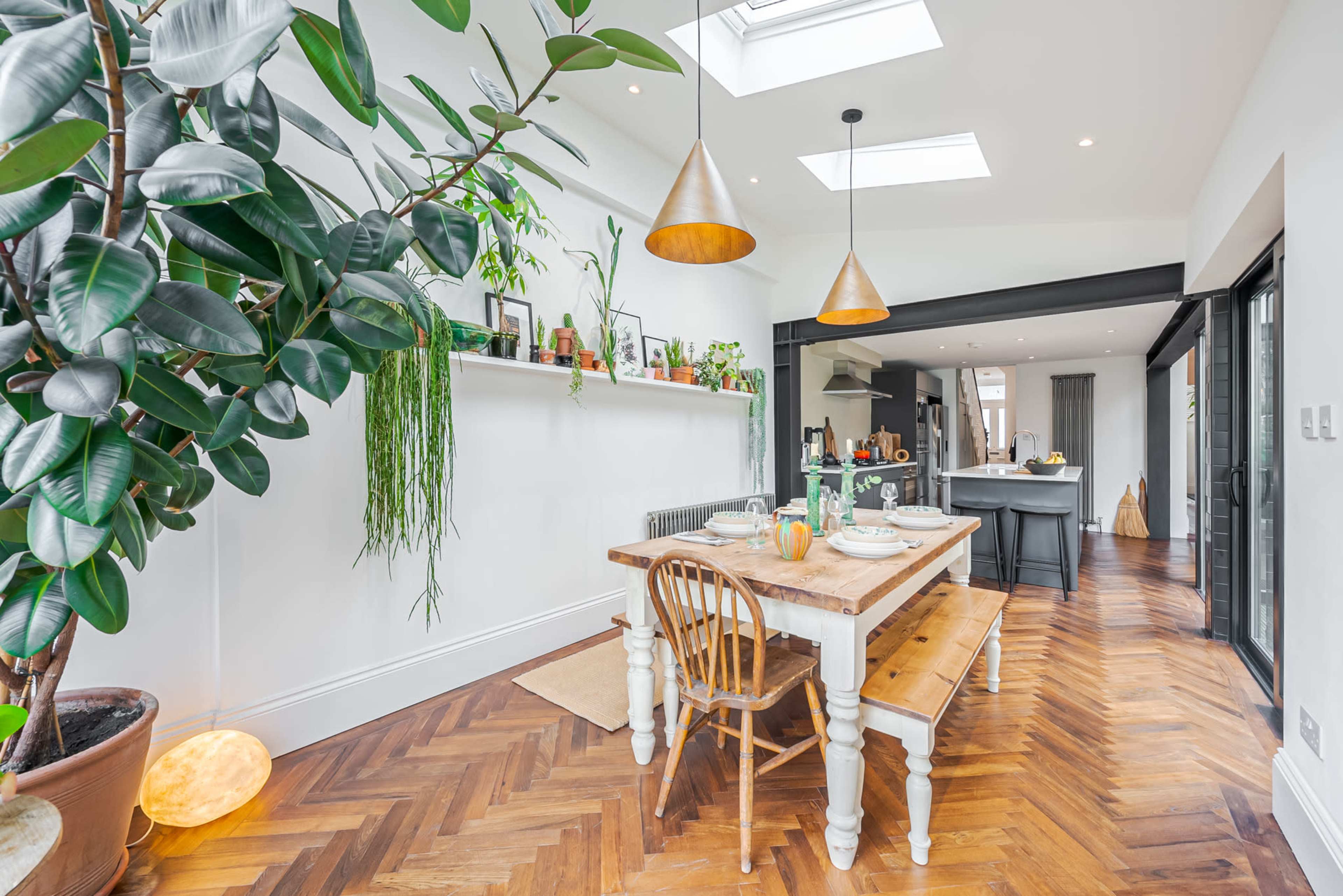 A modern dining area features a wooden table with benches, surrounded by greenery and illuminated by pendant lights.