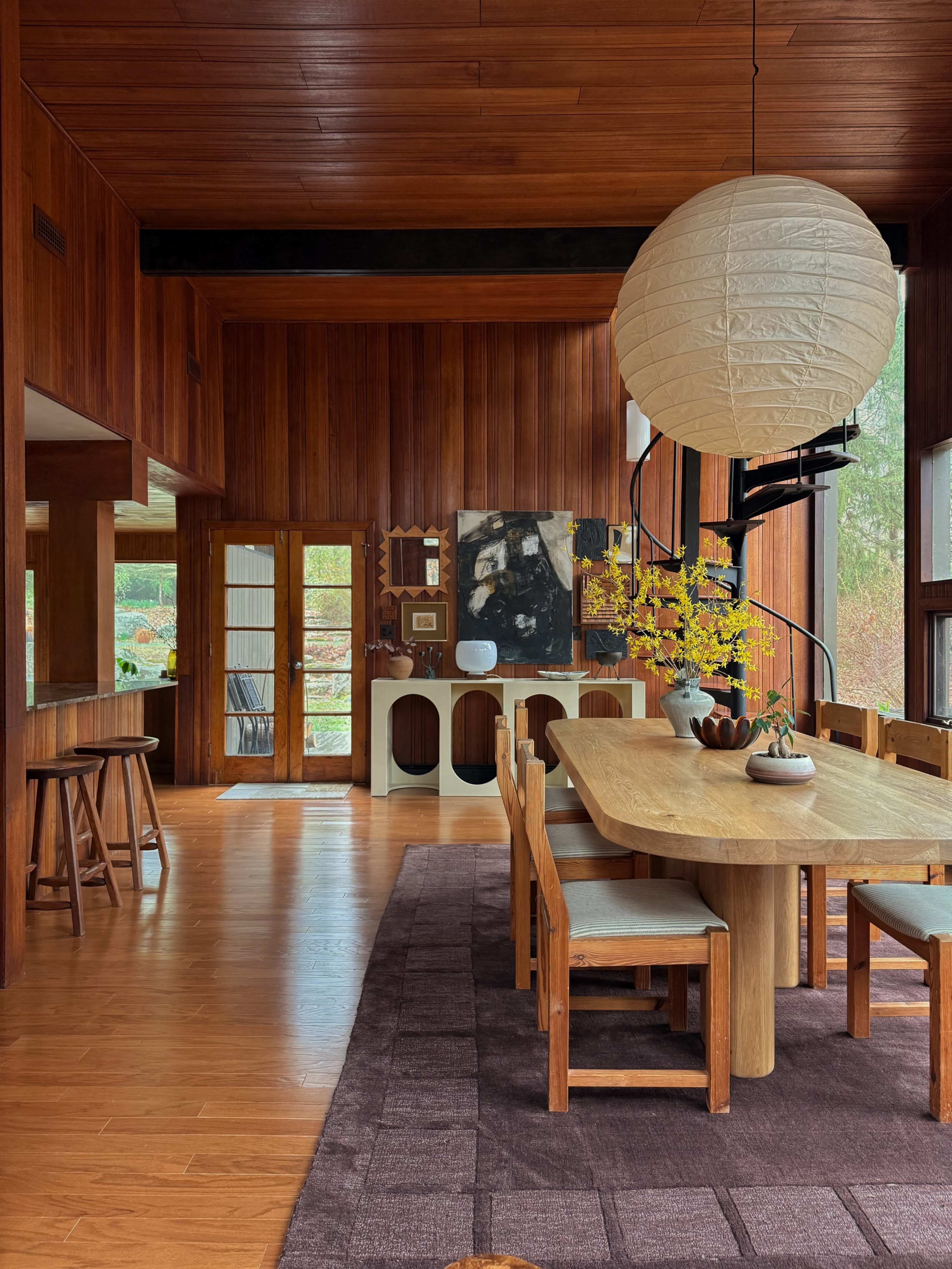 The image shows a spacious dining area featuring wooden furnishings, a large table, and a round pendant light, with large windows allowing natural light to enter.