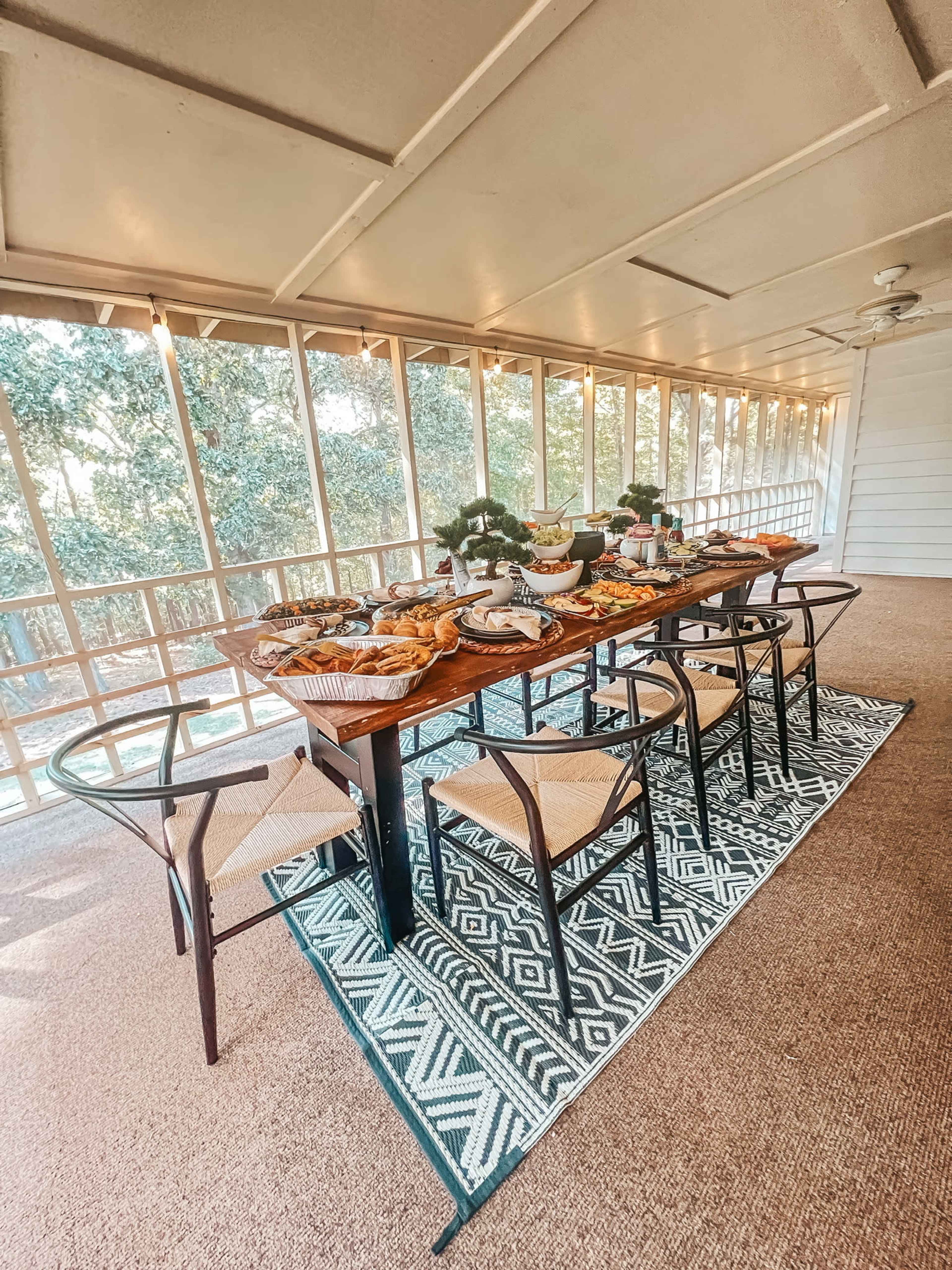 A wooden dining table set with a variety of food surrounded by chairs, located in a sunlit room with large windows.