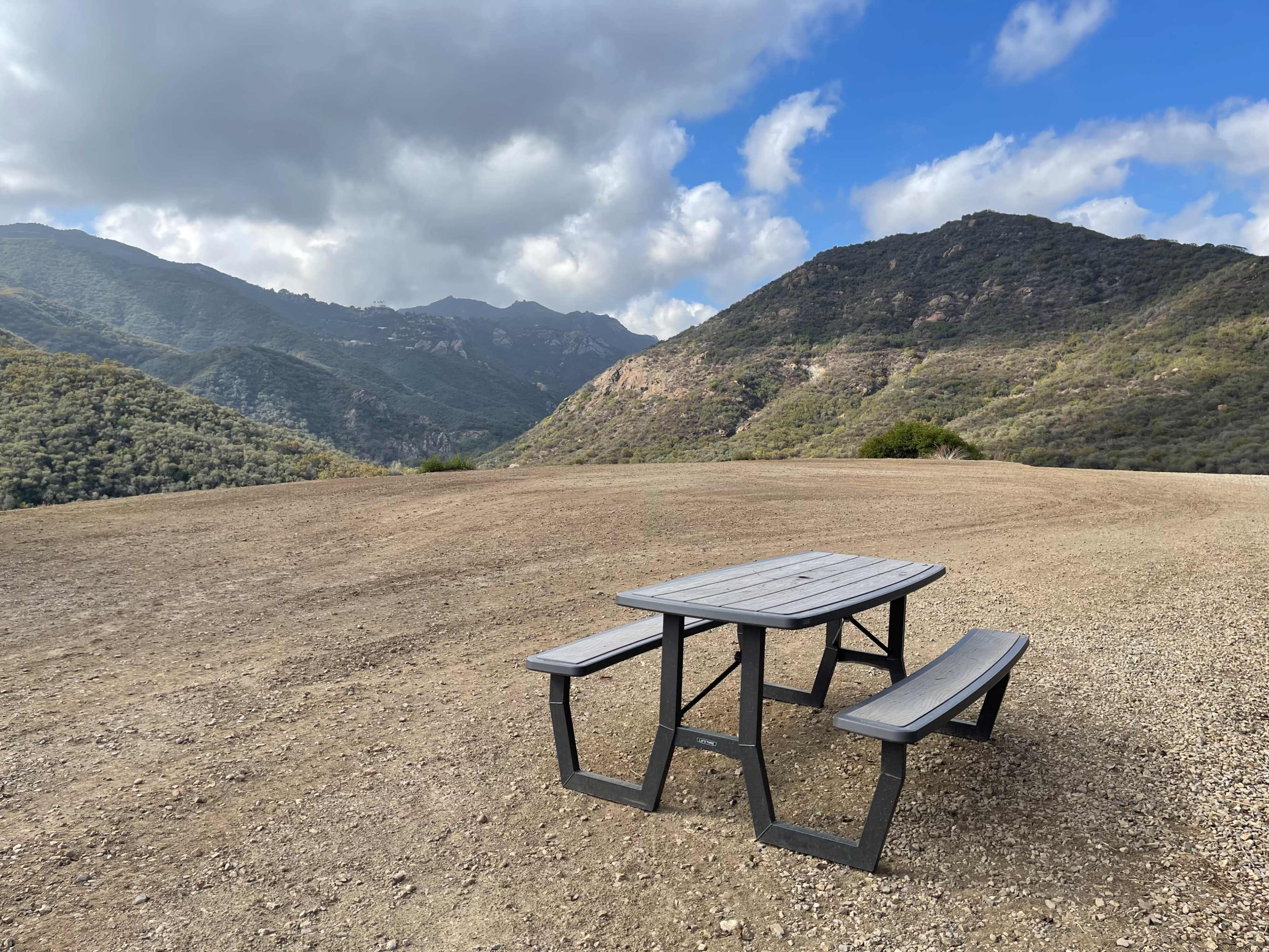 A gray picnic table with attached benches sits on a gravel area overlooking a mountainous landscape under a partly cloudy sky.