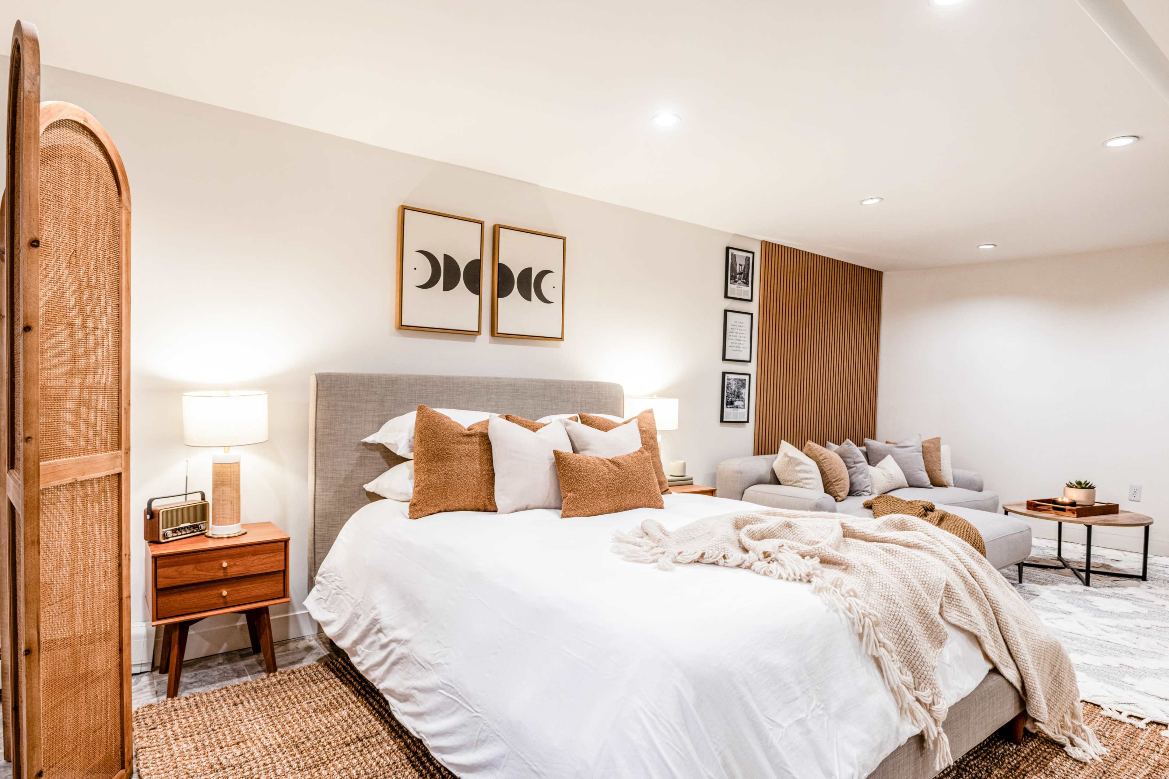 The image shows a cozy bedroom with a large bed, decorative pillows, and an adjacent seating area featuring a couch and a wall adorned with framed art.