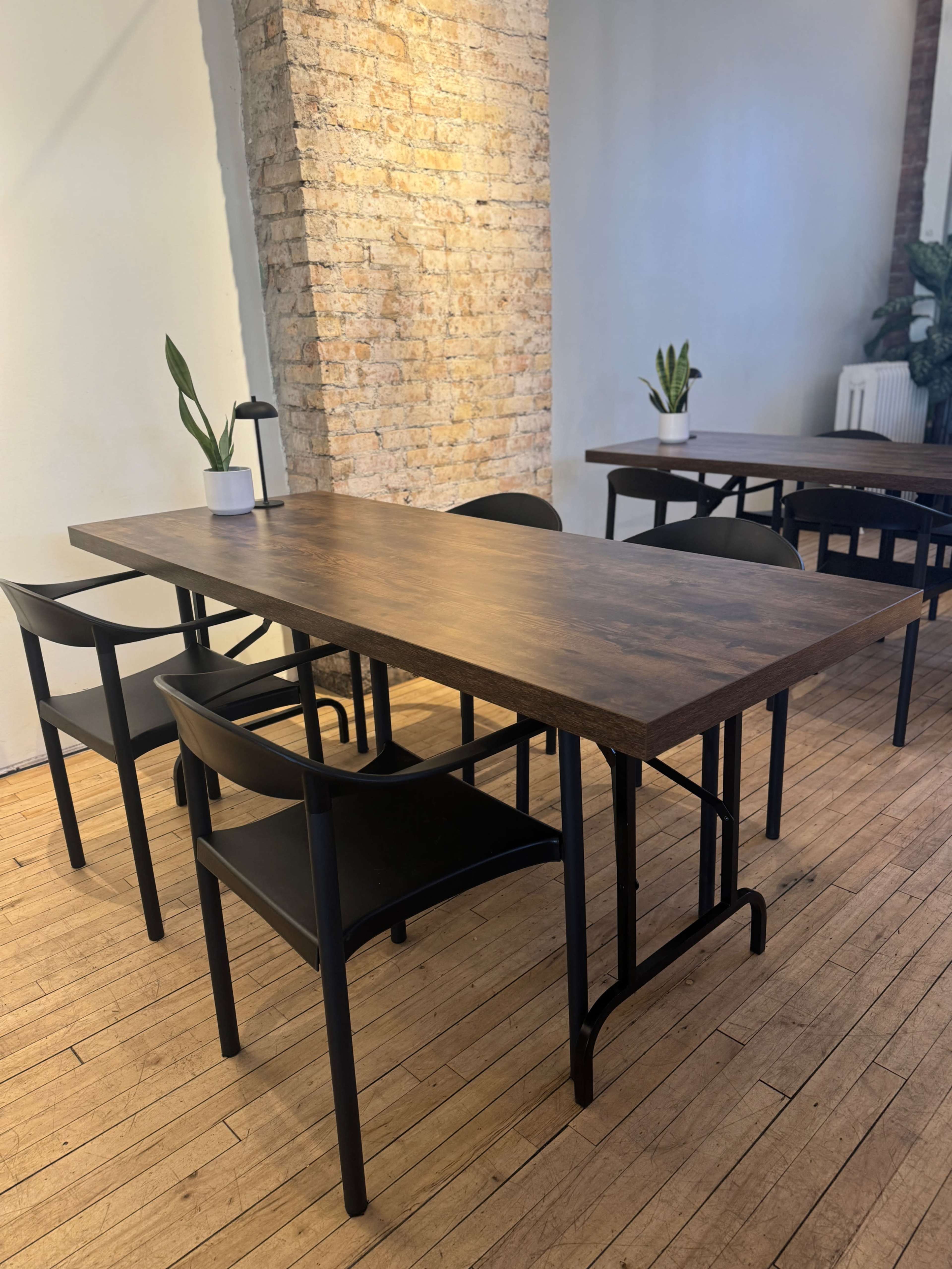 A wooden table with black chairs is arranged in a spacious room featuring a brick wall and minimal decor.
