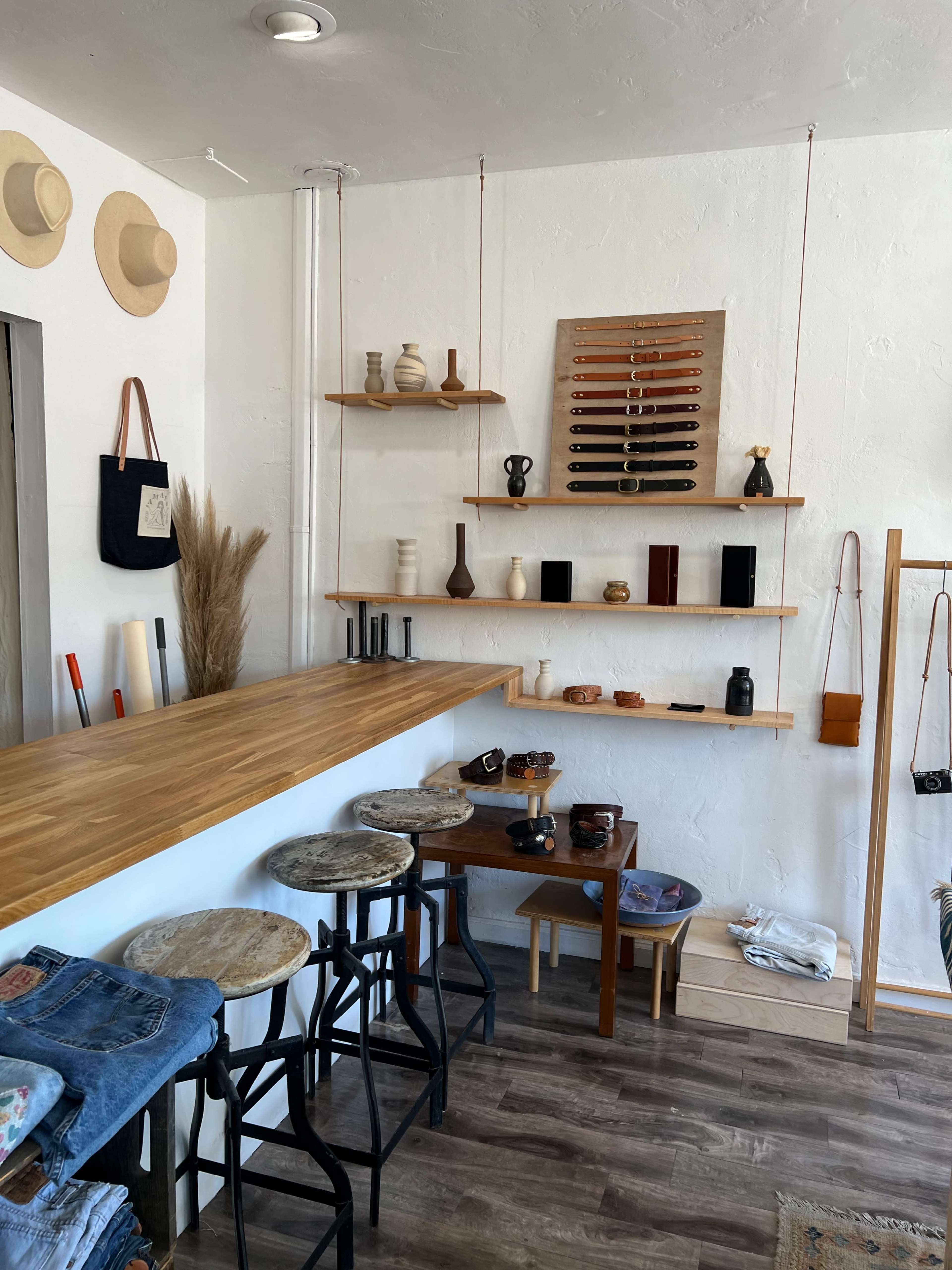 The image shows an interior of a retail space featuring wooden shelves displaying various items, a bar counter with stools, and a section for clothing arranged on the floor.