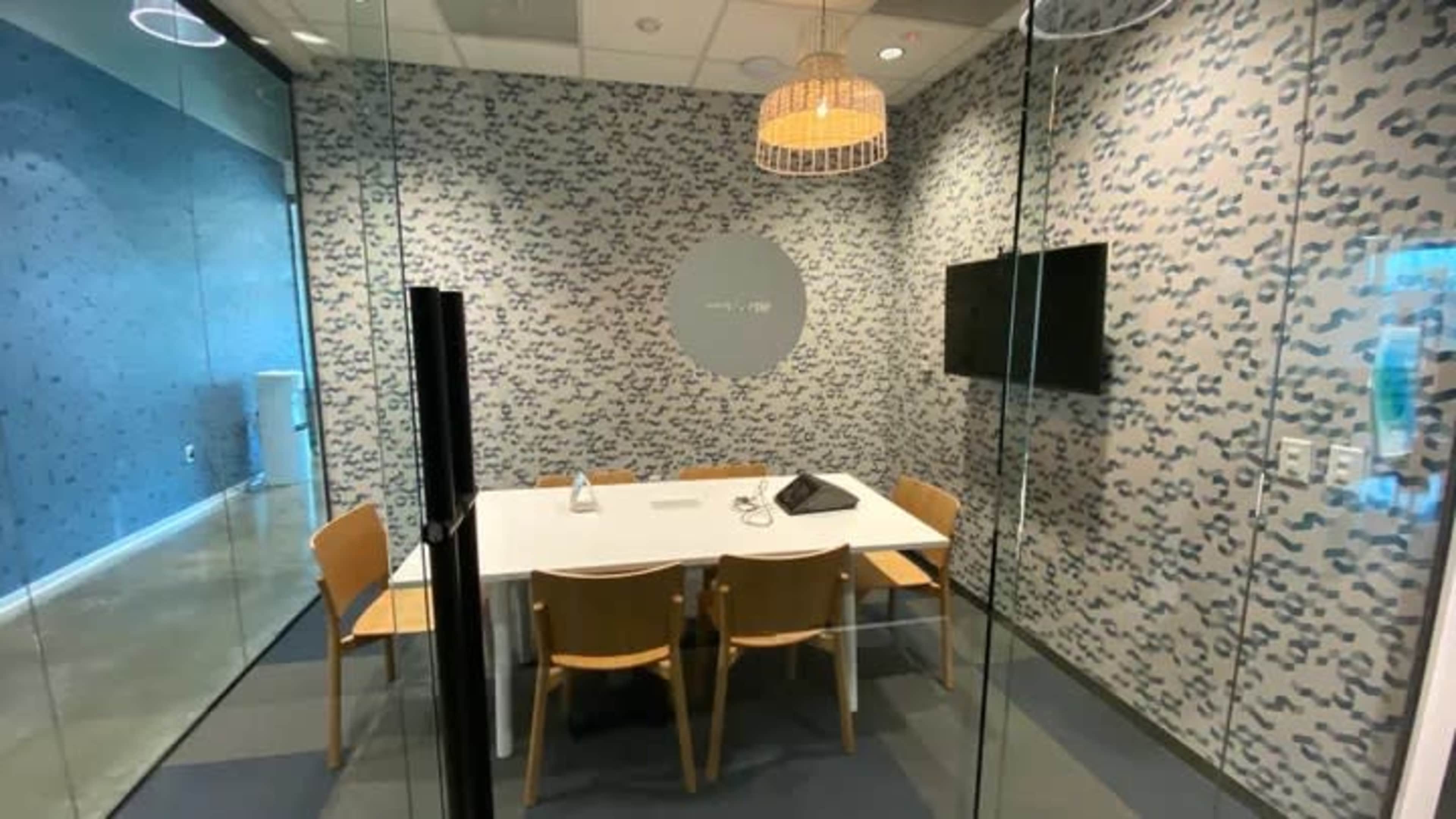 A glass-walled meeting room with a white table, four wooden chairs, a wall-mounted television, and a decorative light fixture overhead.