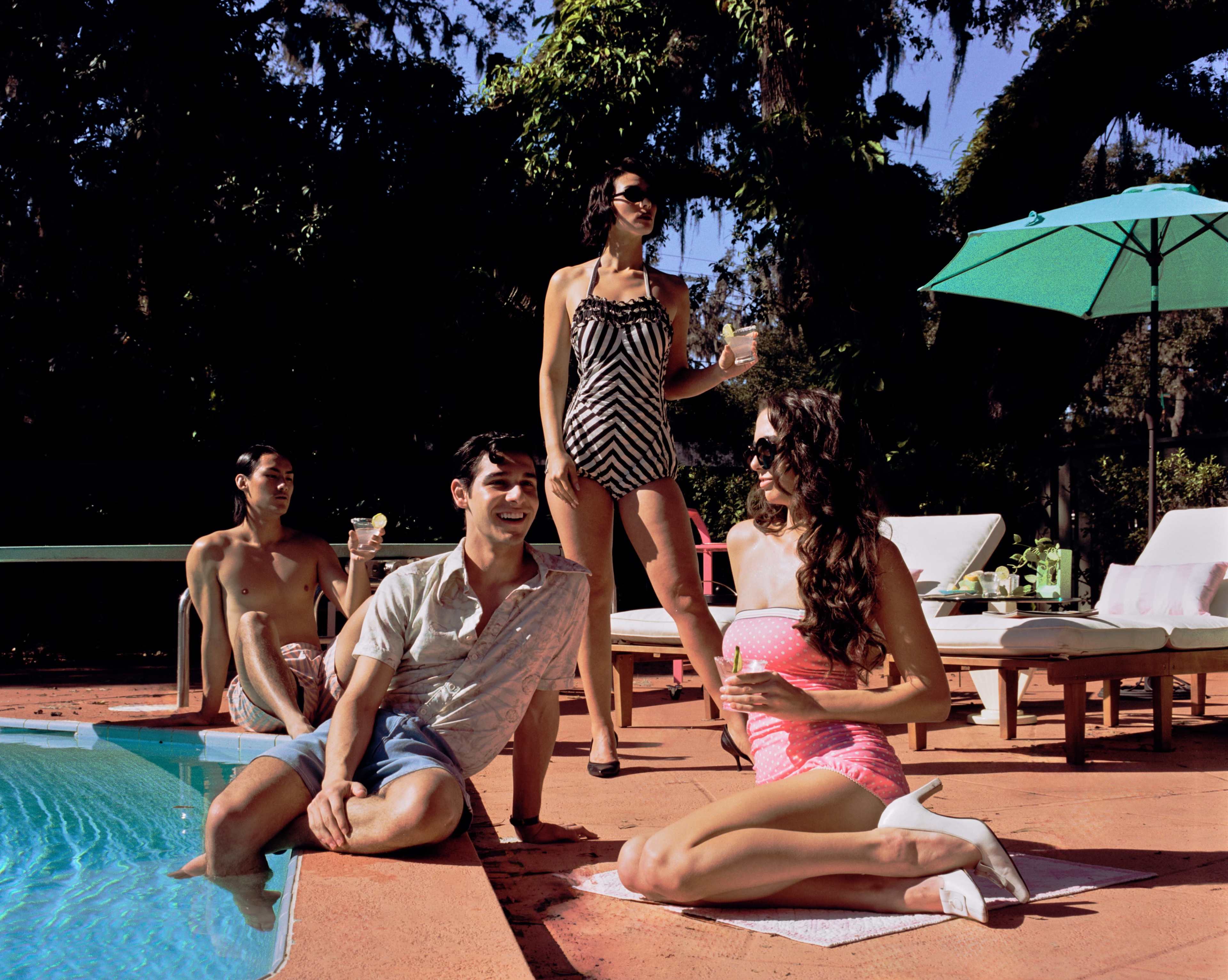 Four people enjoy a sunny day by a poolside, with two sitting on the edge of the pool and two standing nearby.