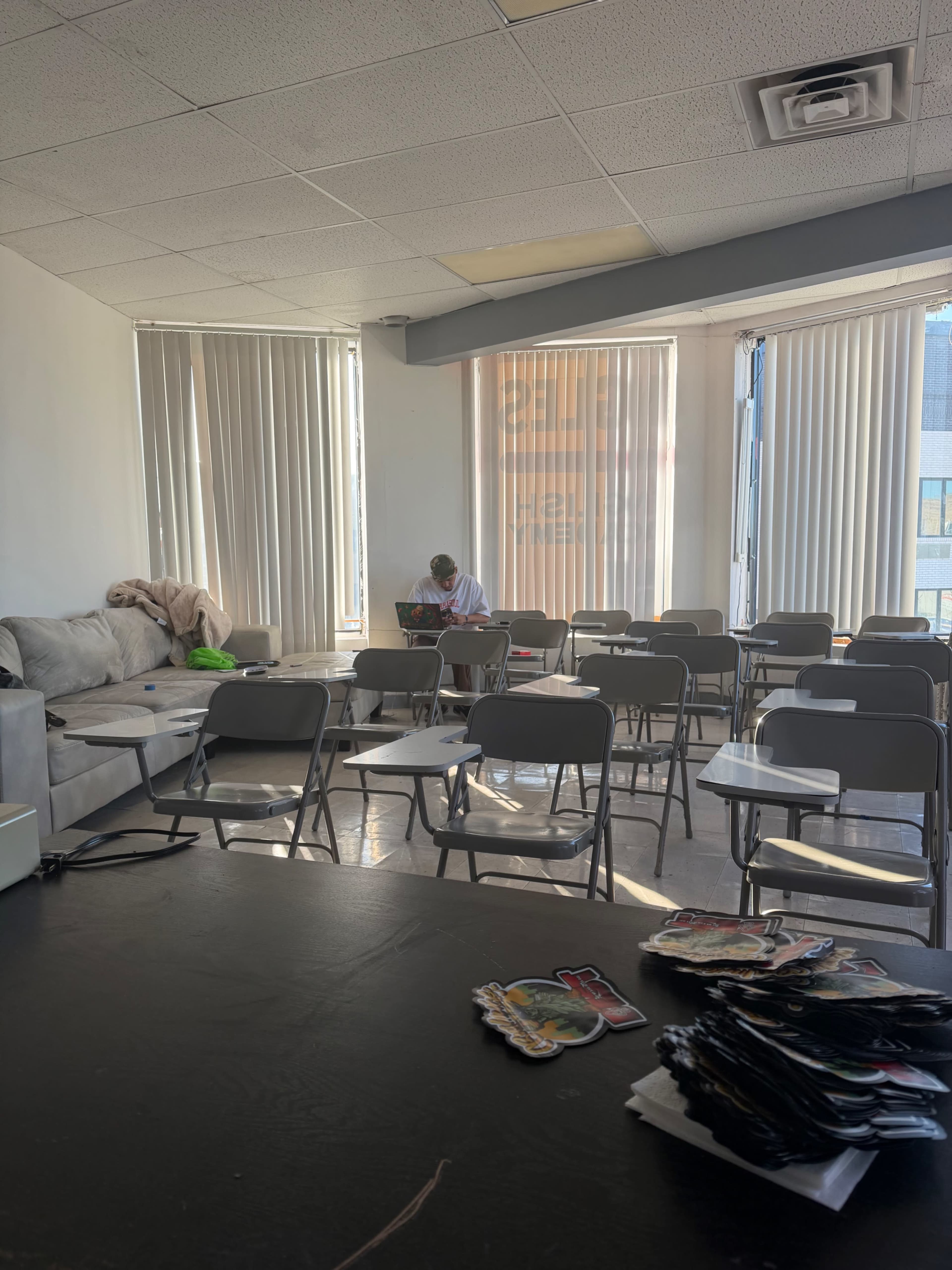 A classroom with several empty chairs and a table, featuring a student focused on a laptop in the background near a window.
